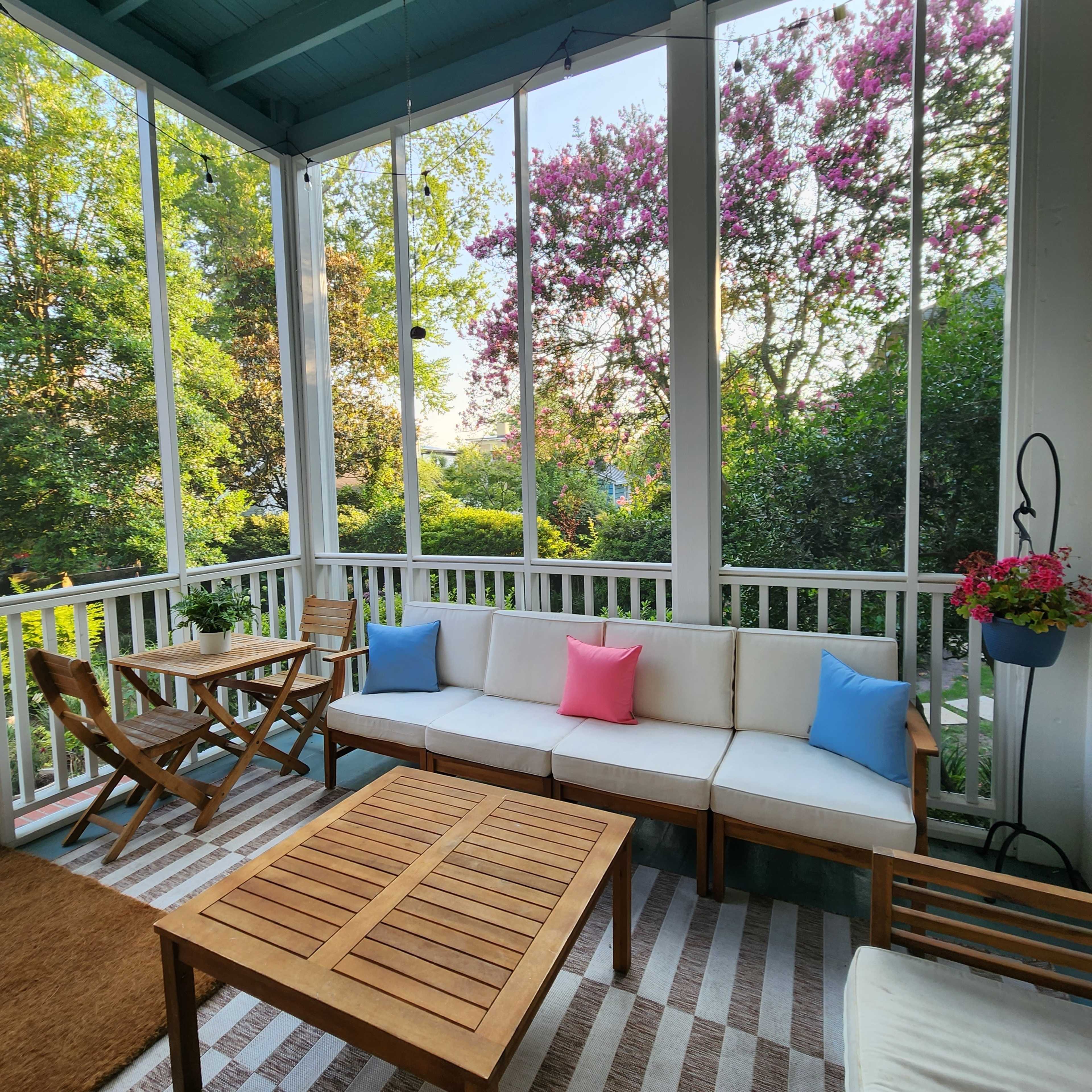 A screened-in porch features a wooden table and chairs, a cushioned sofa with pink and blue pillows, and vibrant greenery outside.
