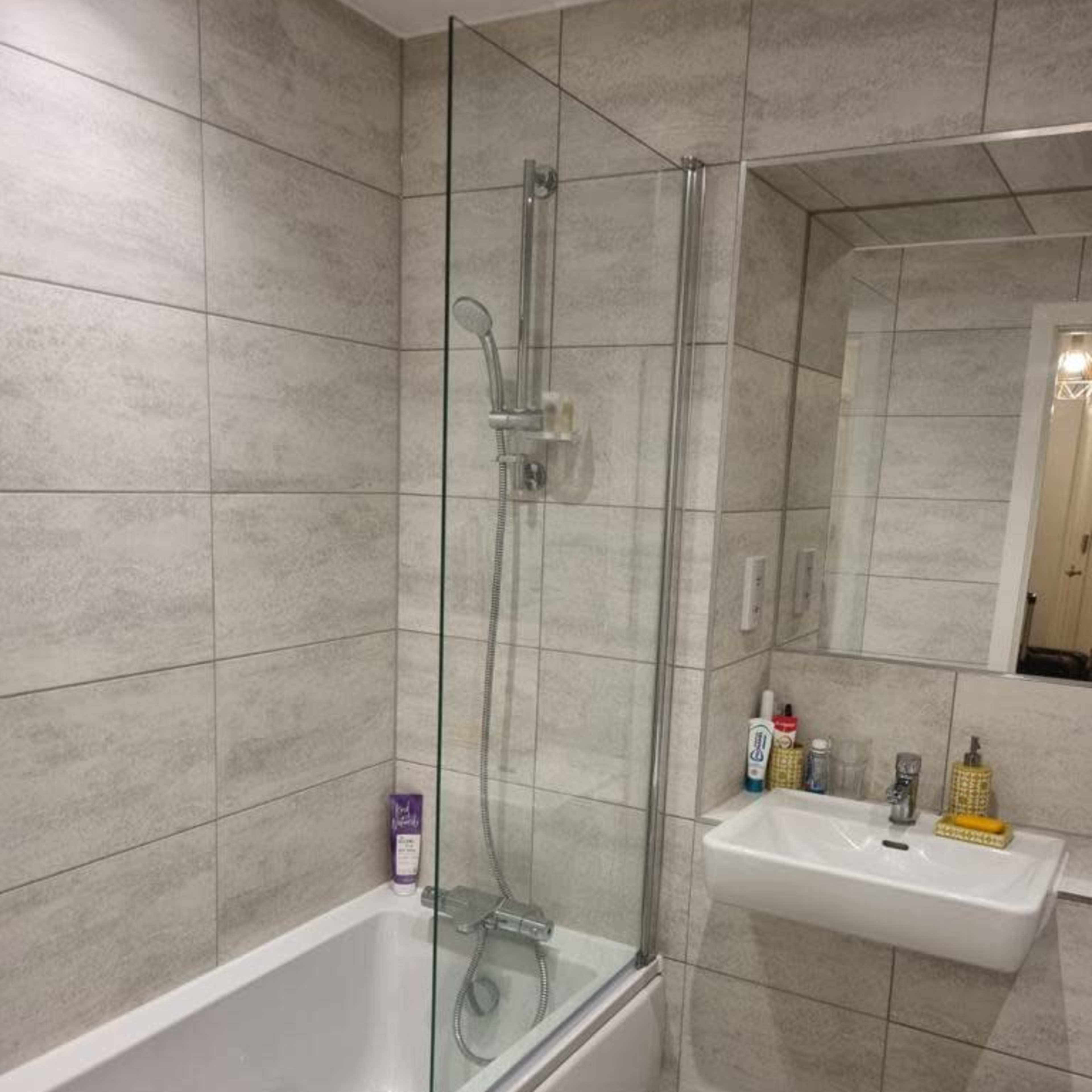A bathroom with a glass shower enclosure over a bathtub, a sink with various toiletries on the counter, and tiled walls and floors.