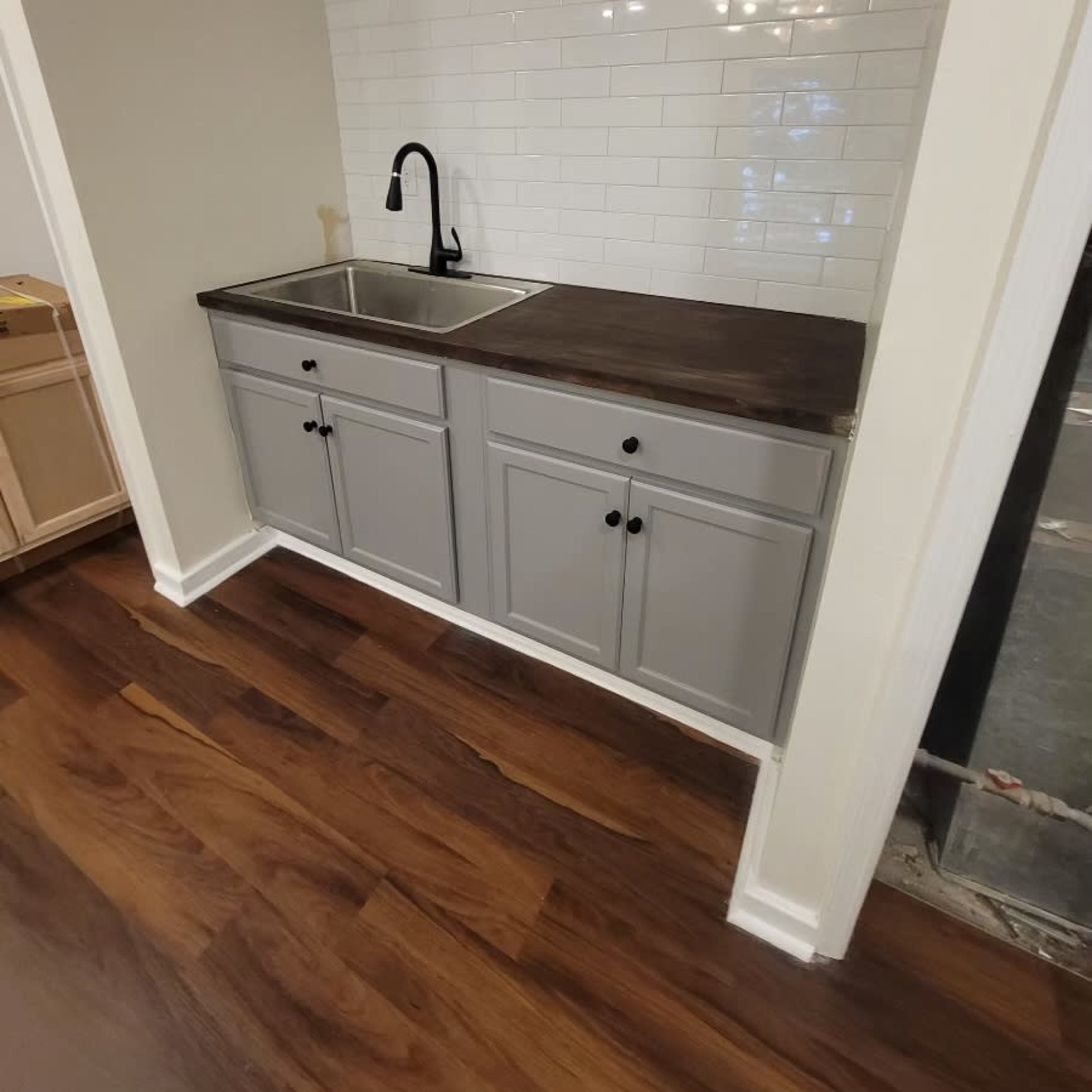 A kitchen area featuring a gray cabinet with a wooden countertop and a single basin sink, set against a white tiled backsplash.