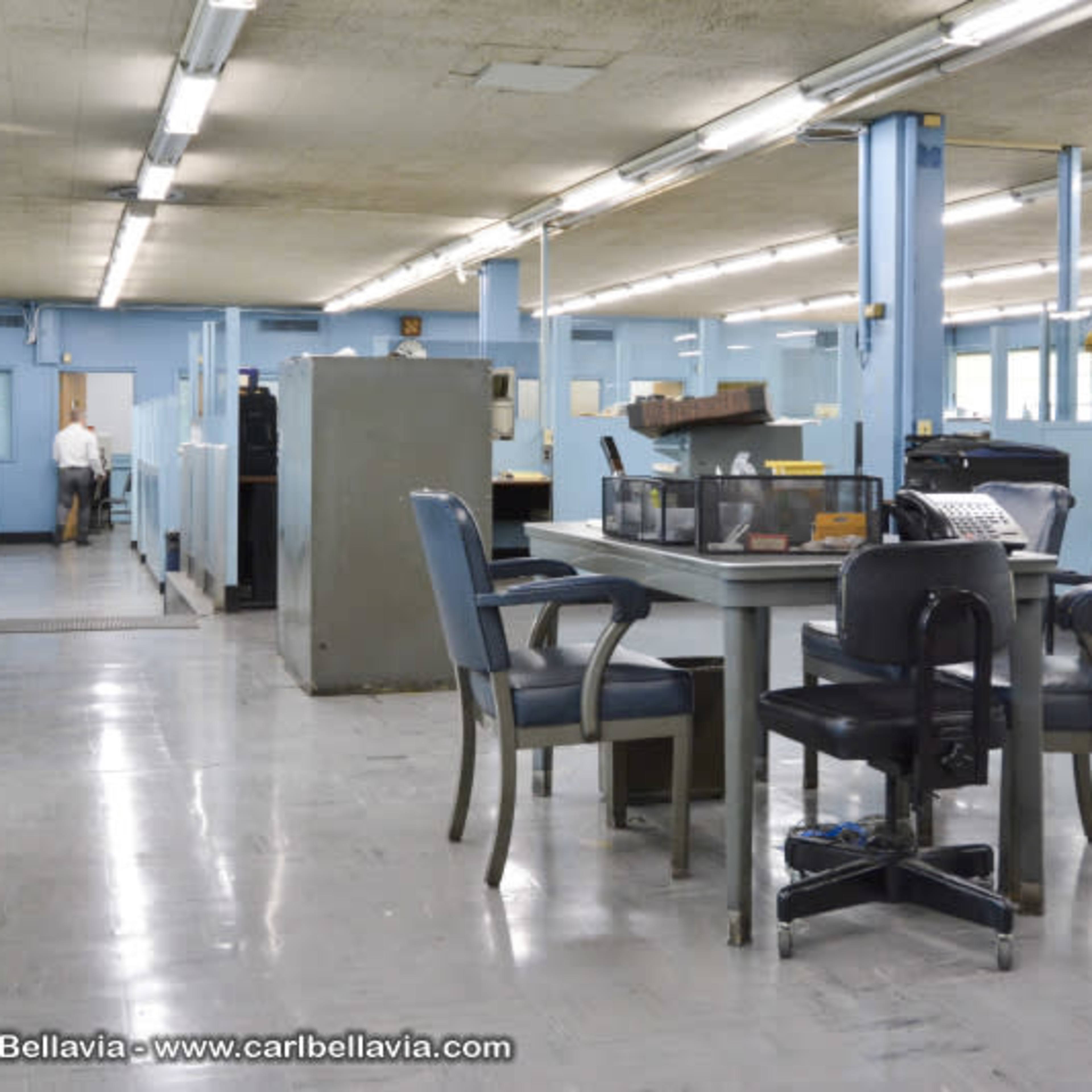 A spacious office interior with blue walls, empty desks, and rows of chairs.