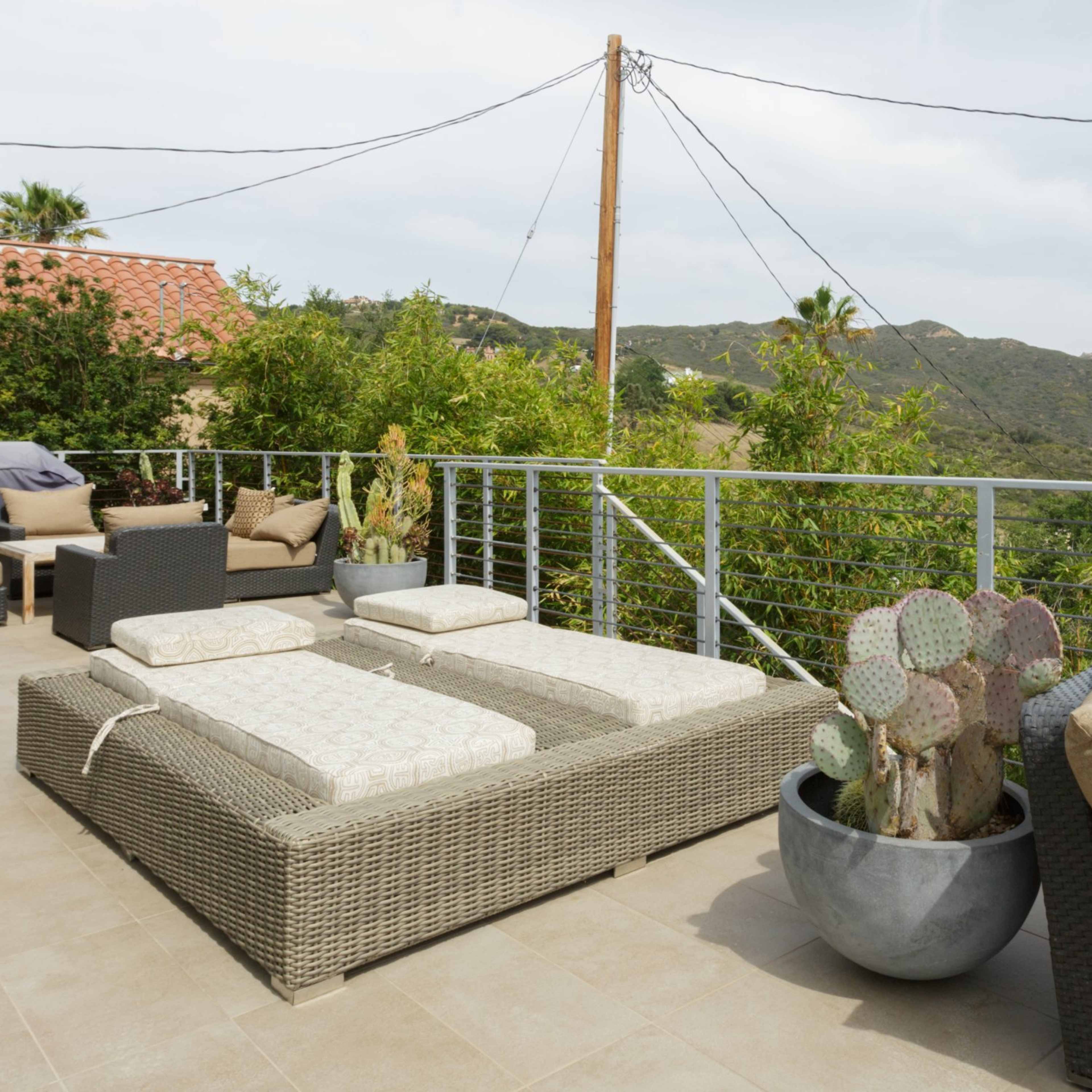 A terrace with lounge beds and a comfortable chair overlooks a mountainous landscape, surrounded by greenery and a cactus in a planter.