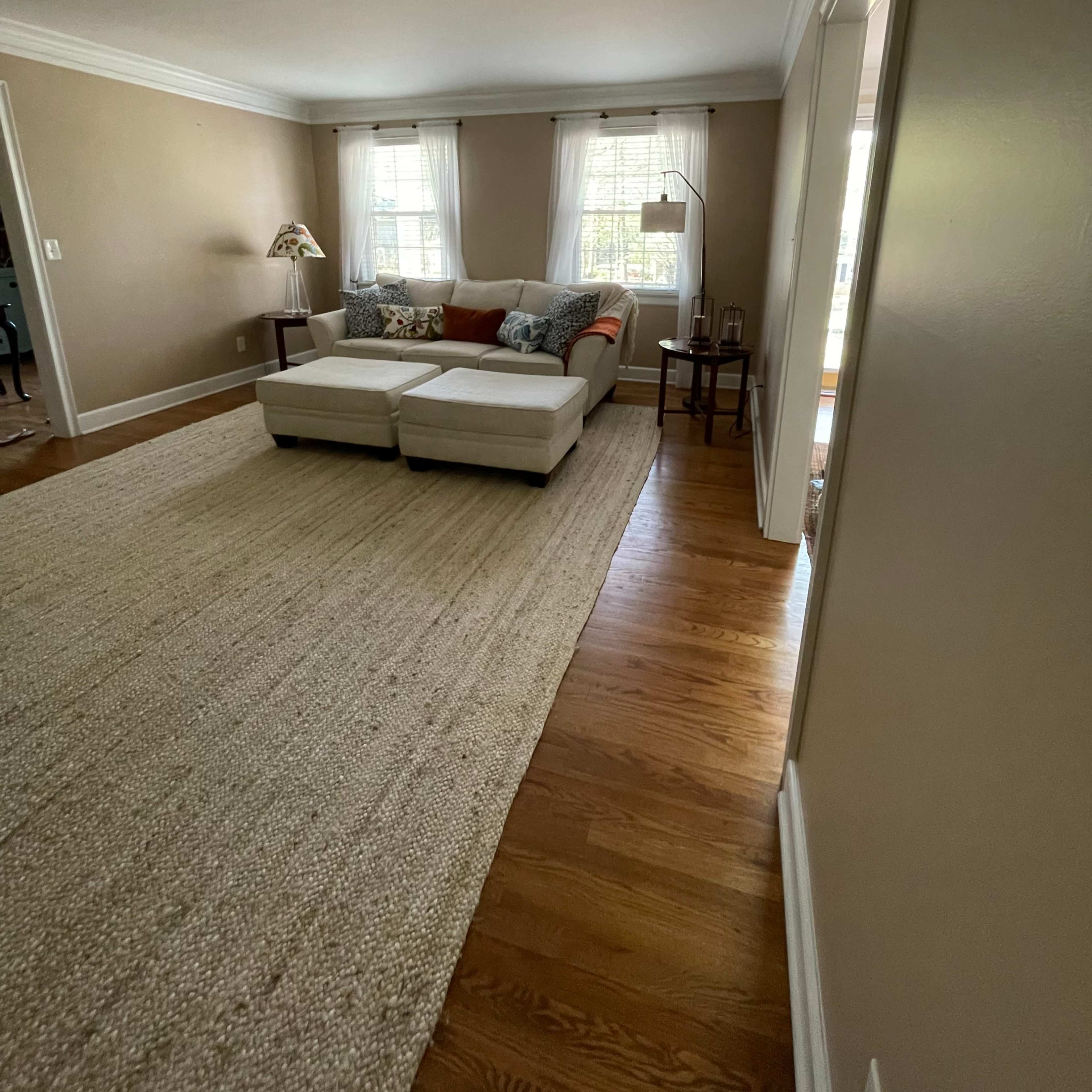 A light-filled living room features a white sectional sofa, a woven area rug, and windows draped with sheer curtains.