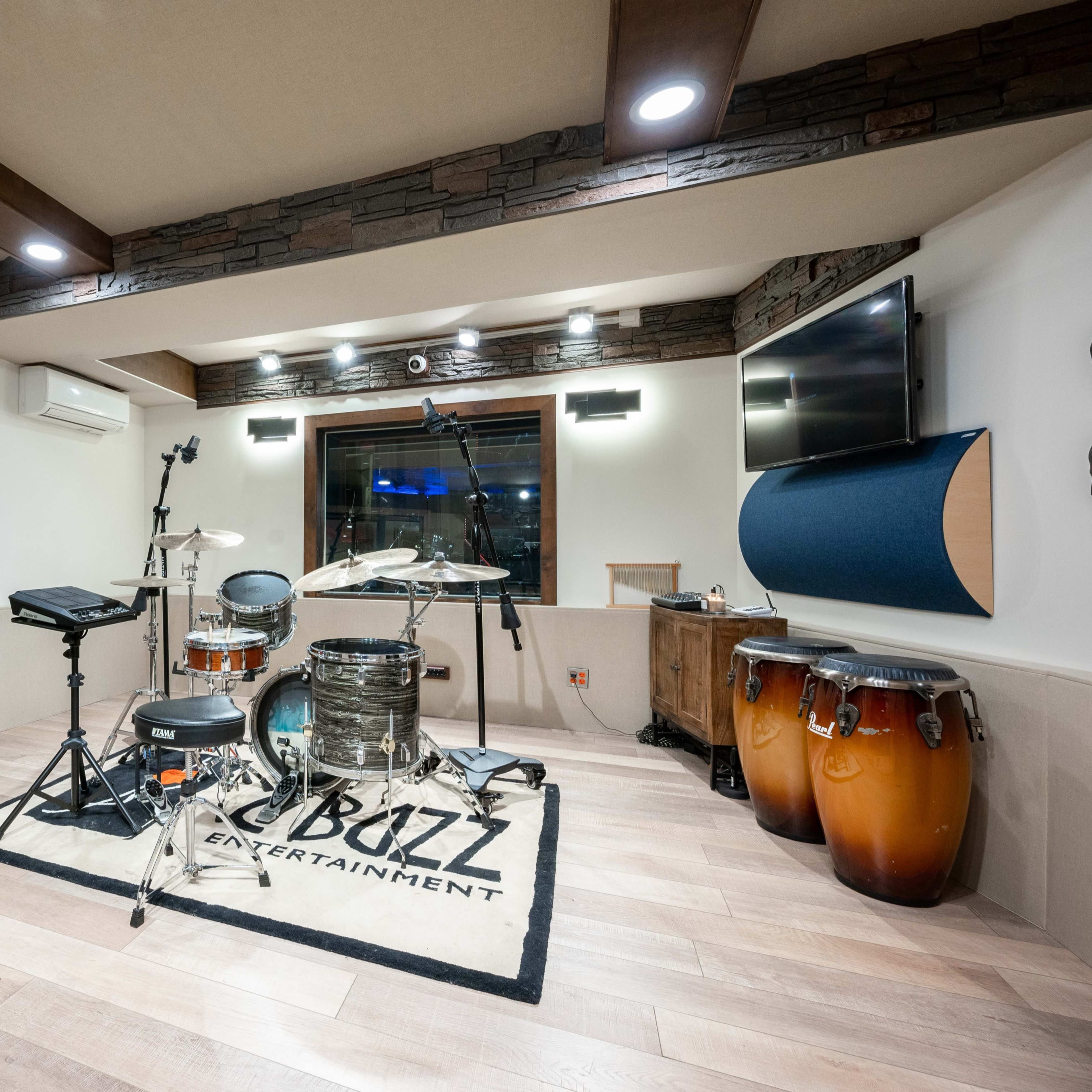 A well-equipped music studio featuring a drum set, conga drums, and various electronic music instruments arranged on a wooden floor with acoustic panels on the walls.