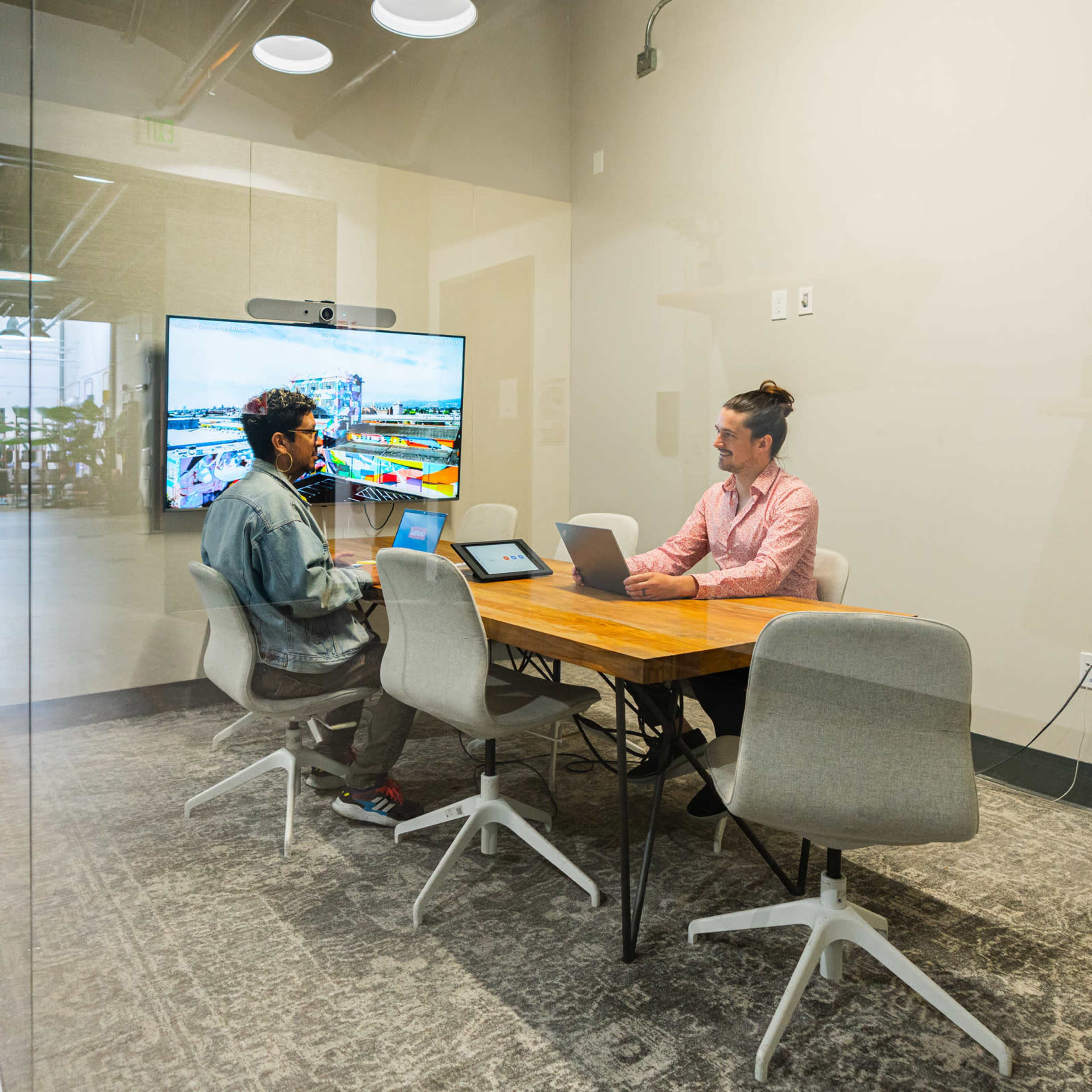 Two people sit at a table in a glass-walled conference room, working on laptops with a large screen displaying a colorful presentation in the background.