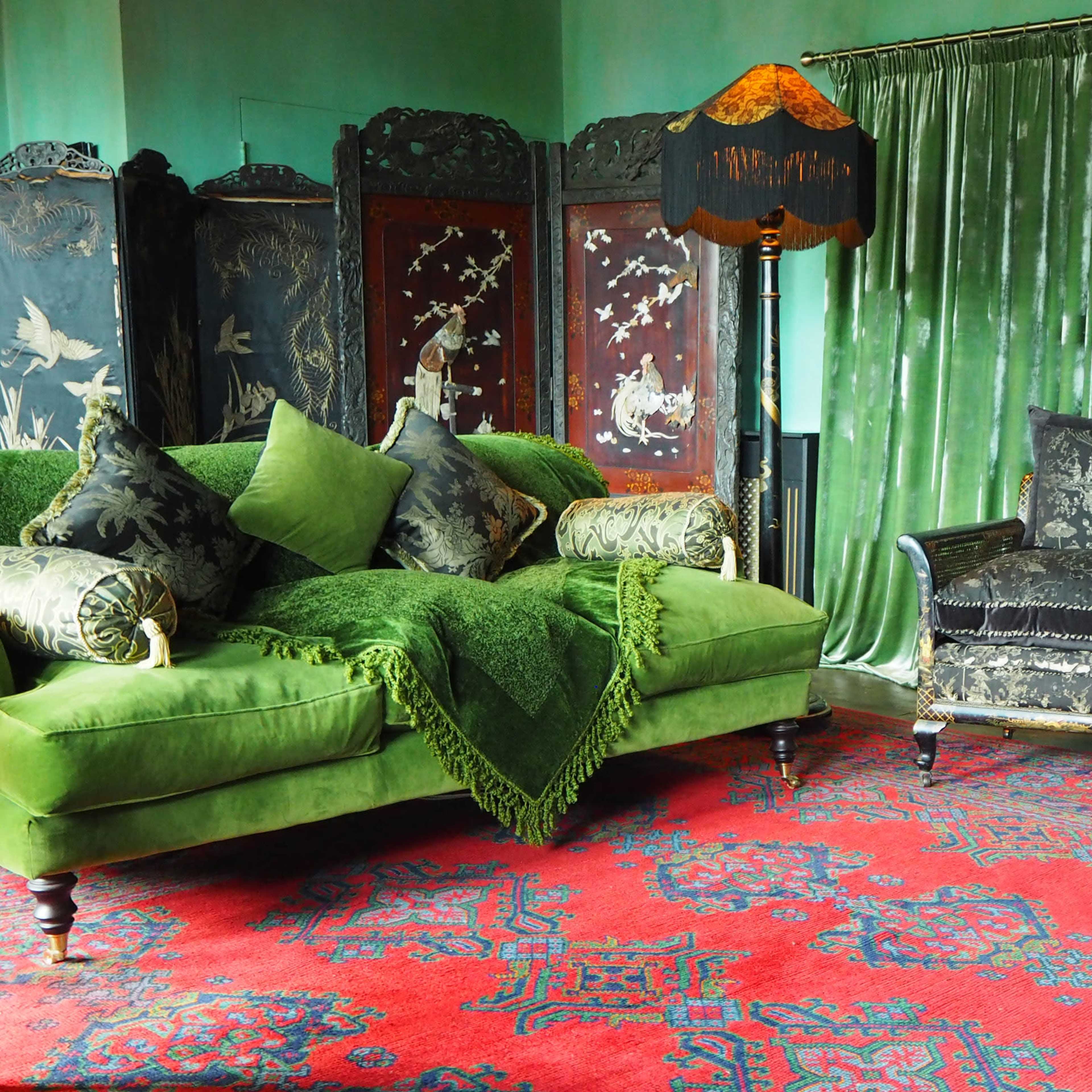 A green sofa adorned with decorative pillows, a matching armchair, and a dark red patterned carpet, set against a backdrop of green curtains and ornate room dividers.