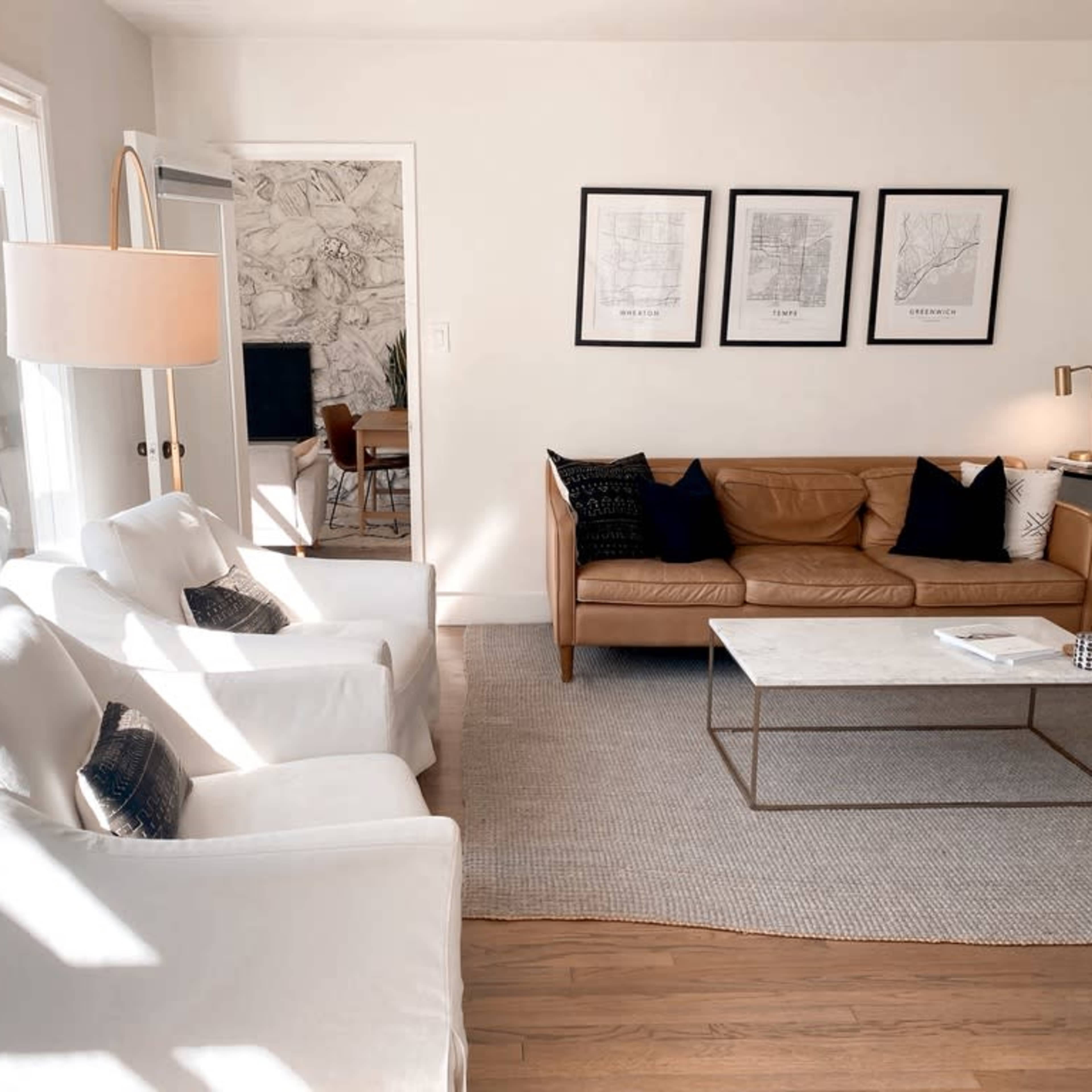 A bright living room features a leather sofa, two white armchairs, a marble coffee table, and framed artwork on the walls.