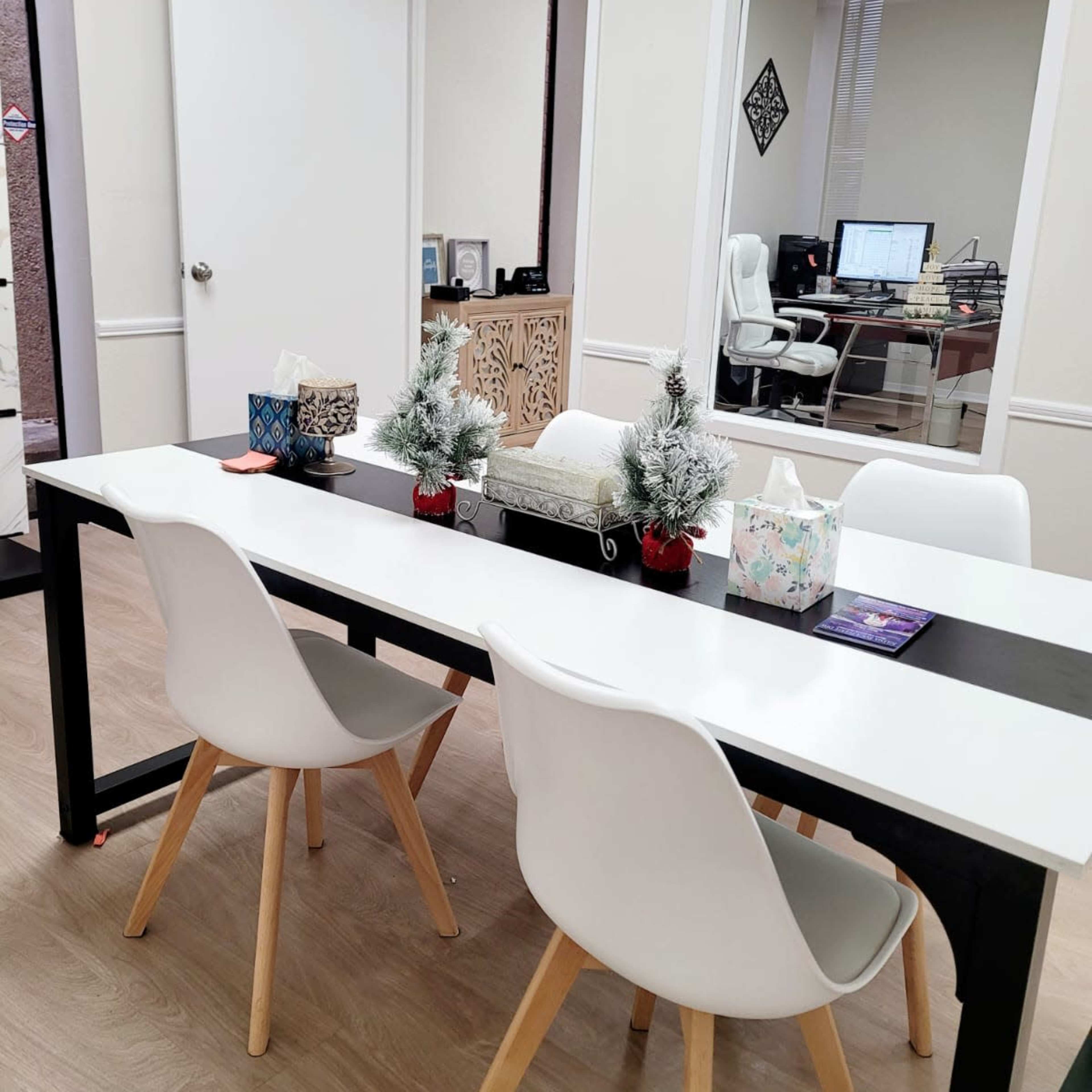 A modern office space features a rectangular table surrounded by four white chairs, adorned with small evergreen decorations and tissue boxes, with a separate workspace visible through a glass window.