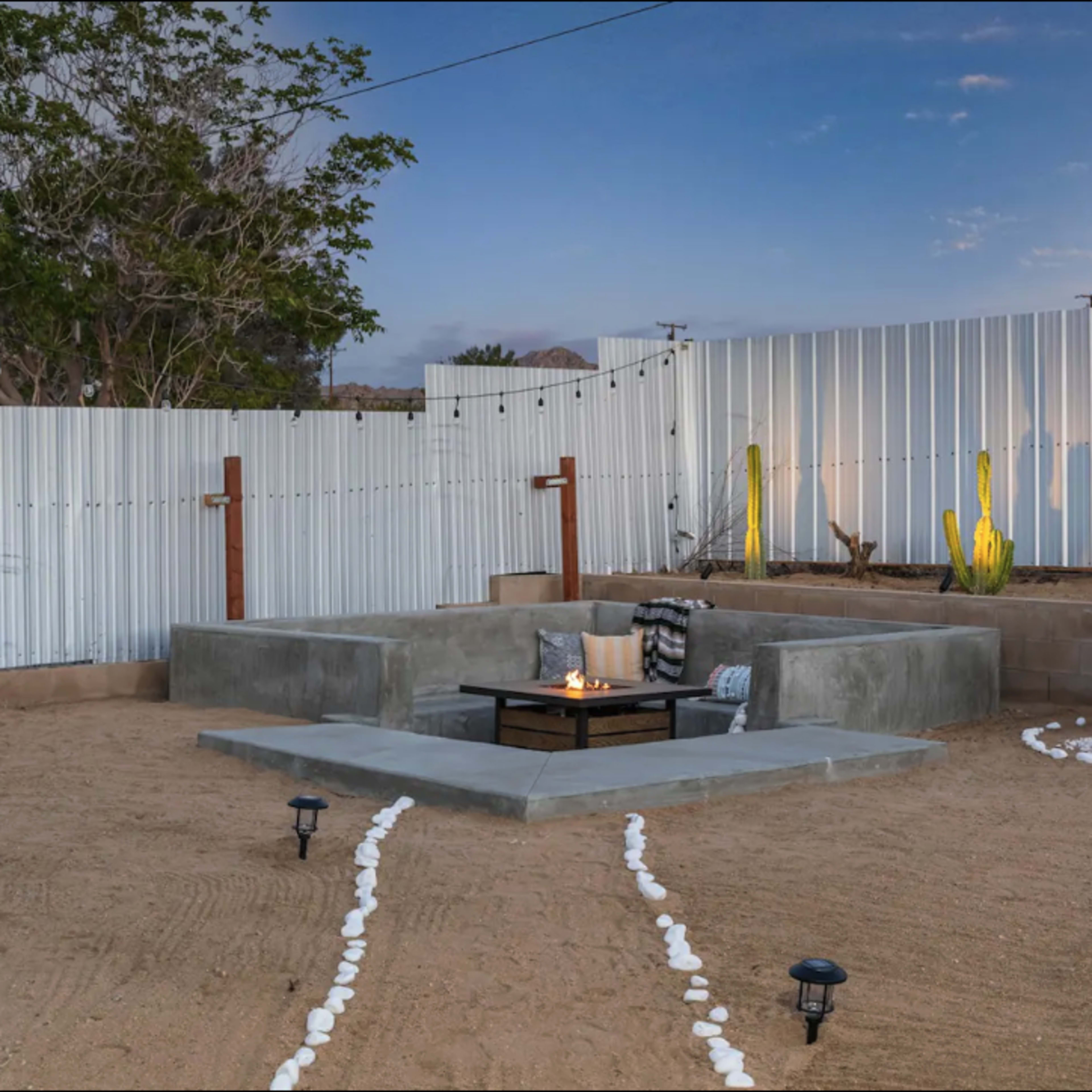 A minimalist backyard space with a raised seating area around a small fire pit, bordered by gravel and illuminated by outdoor lights.