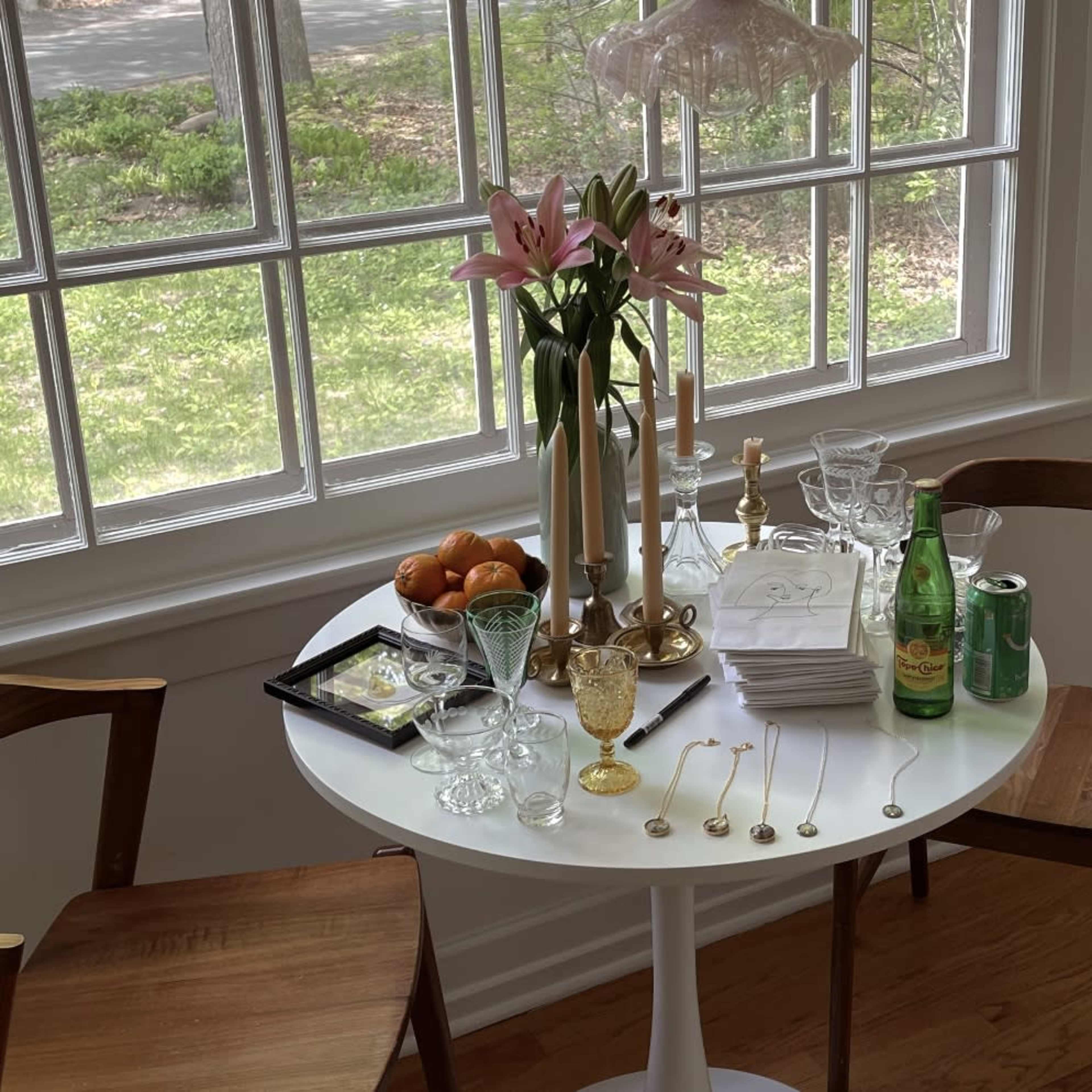 A round table is set with a vase of lilies, candles, fruit, glassware, and two chairs in a bright room with large windows.