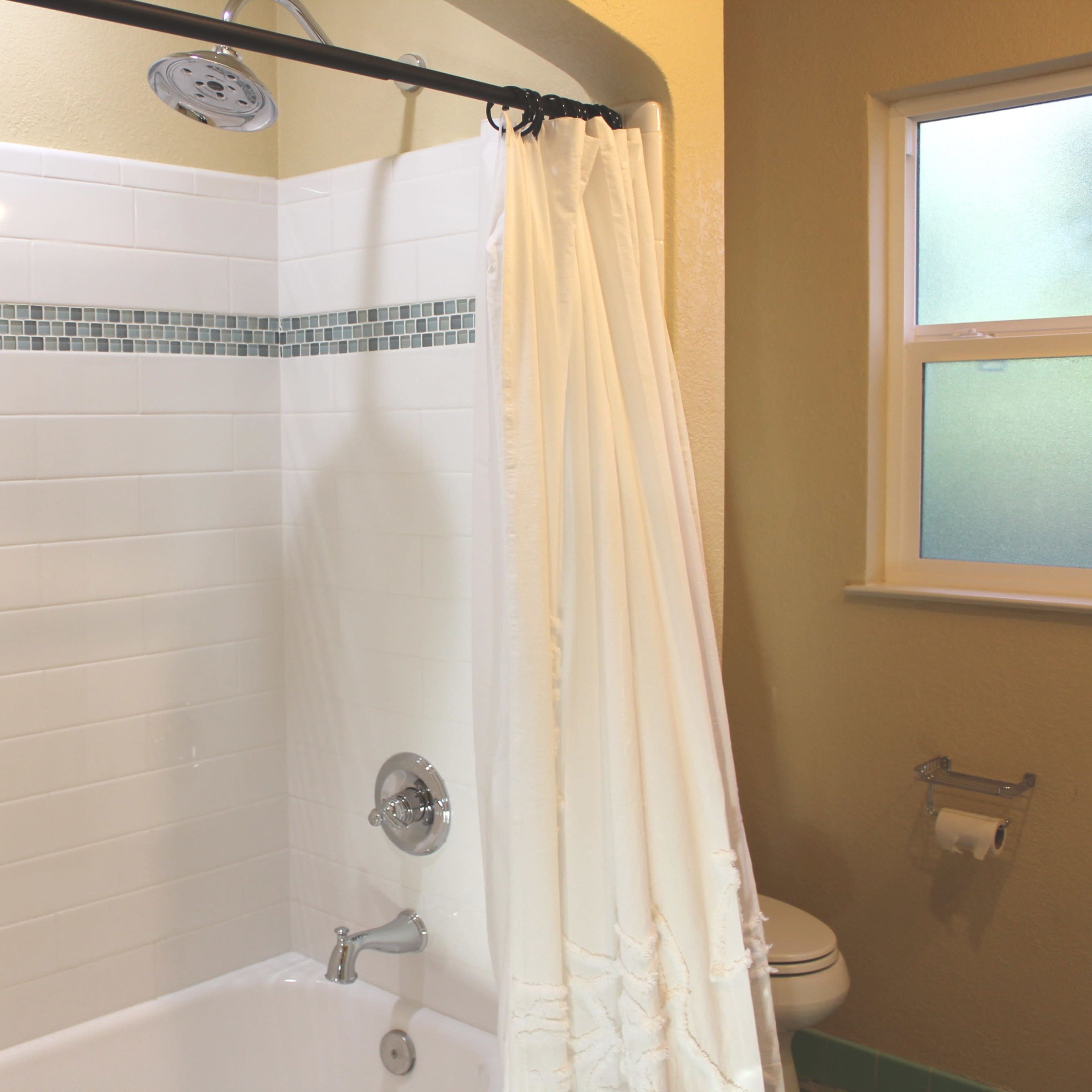 A tiled shower area with a white curtain, a showerhead mounted on a chrome rod, and a toilet in the background.