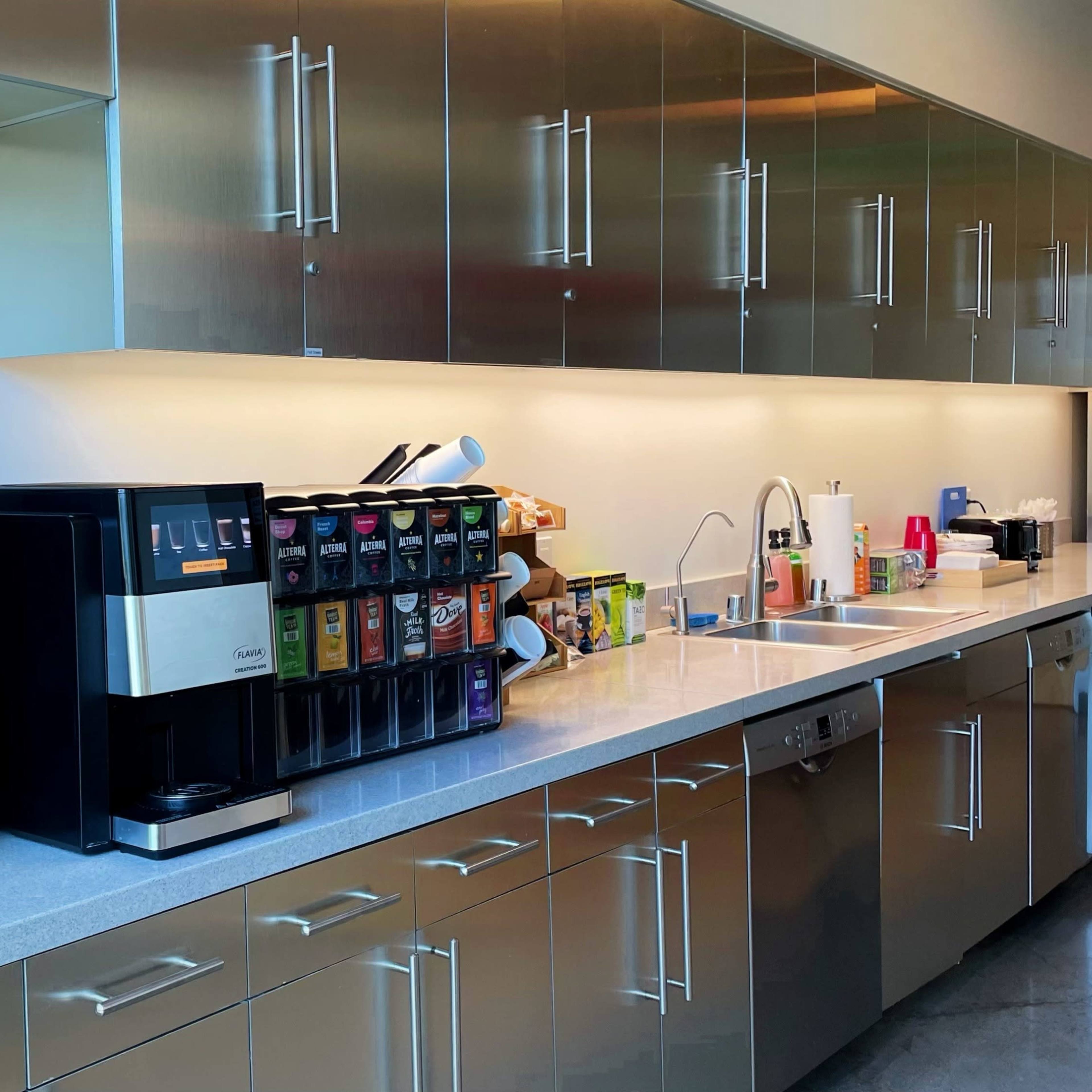 A modern kitchen features stainless steel cabinets, a coffee maker, a drink dispenser, and a sink along a long countertop.