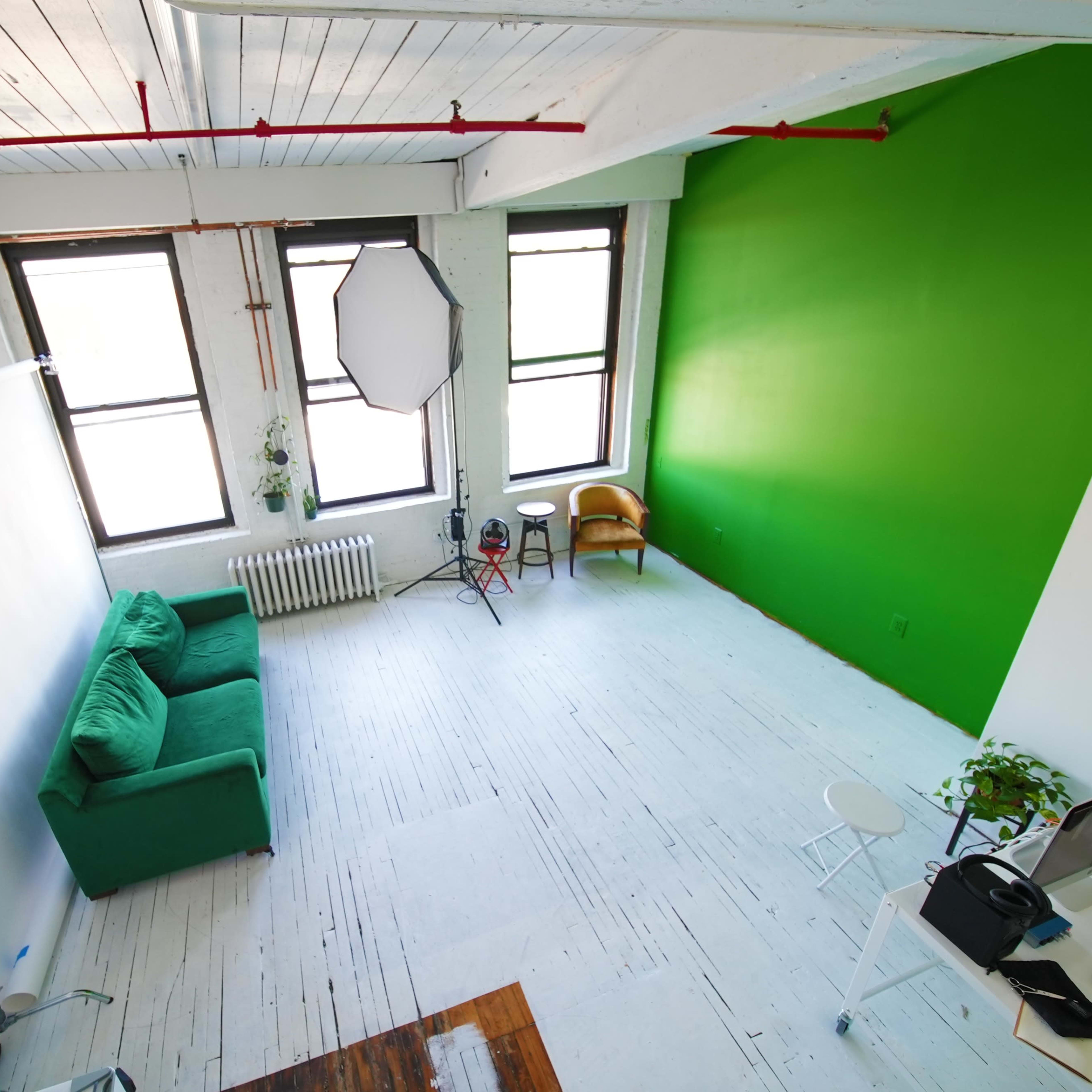 A bright, spacious studio with a green accent wall, a green sofa, a white workspace, and a light setup.