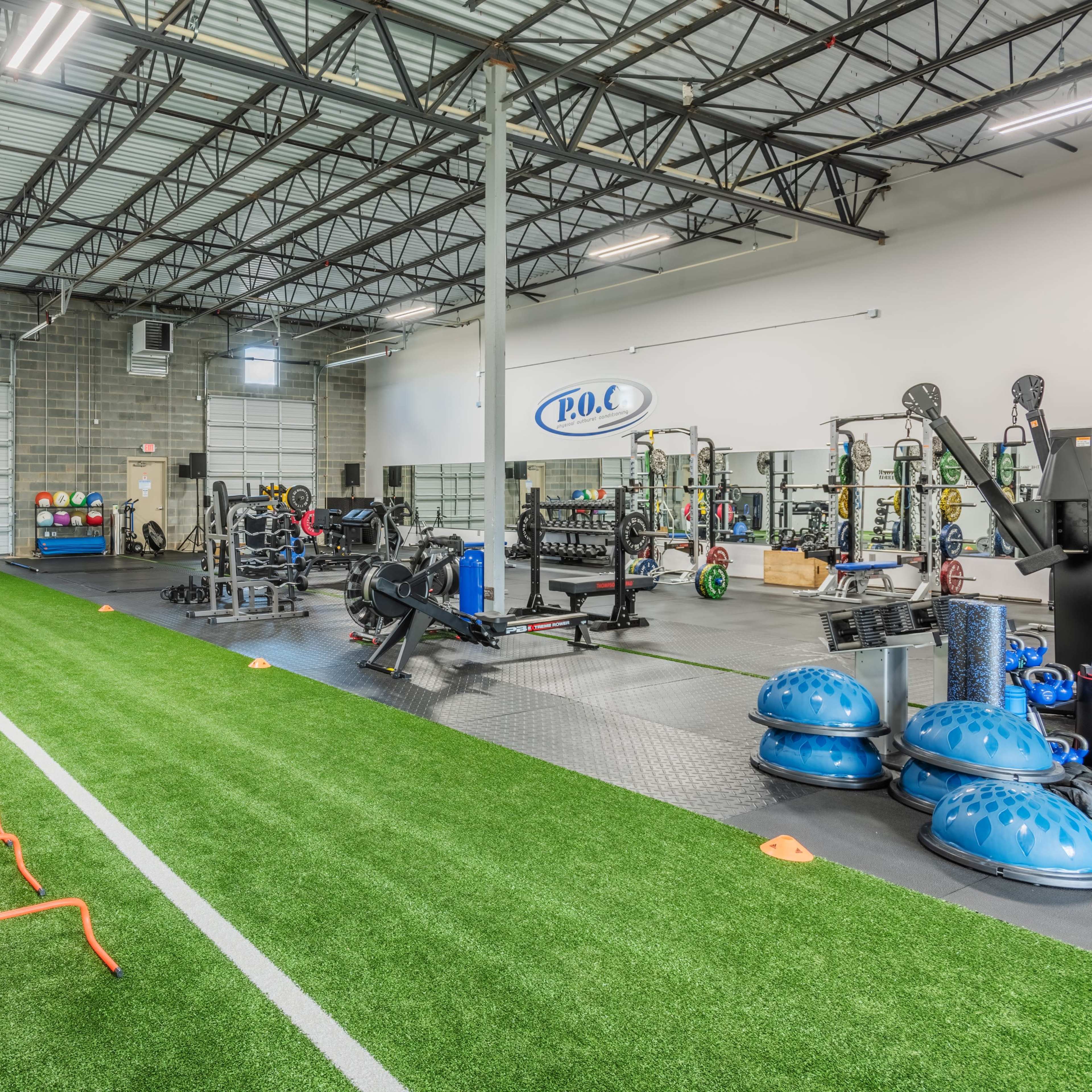 A spacious gym featuring various exercise equipment, a turf area with agility hurdles, and a weightlifting section.