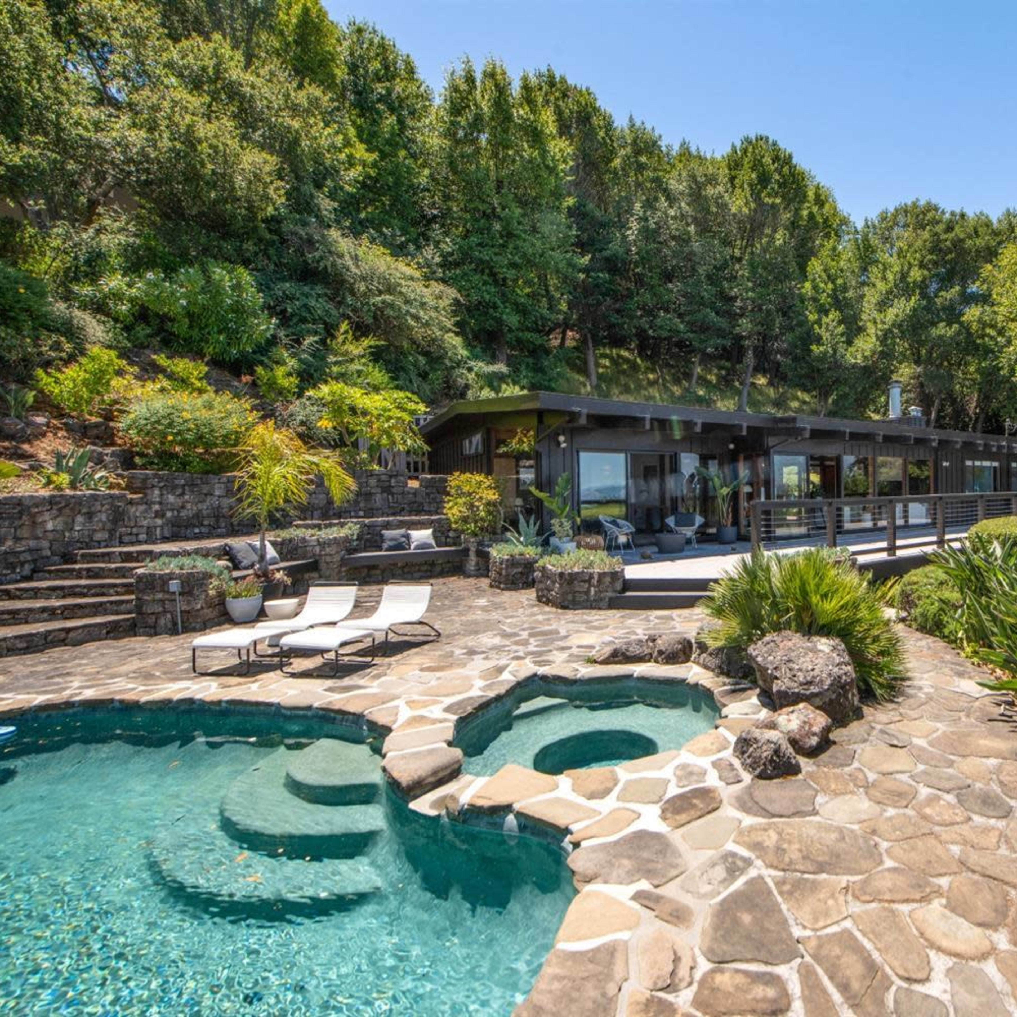 A modern house with large windows surrounded by lush greenery, featuring a stone patio and a swimming pool in the foreground.