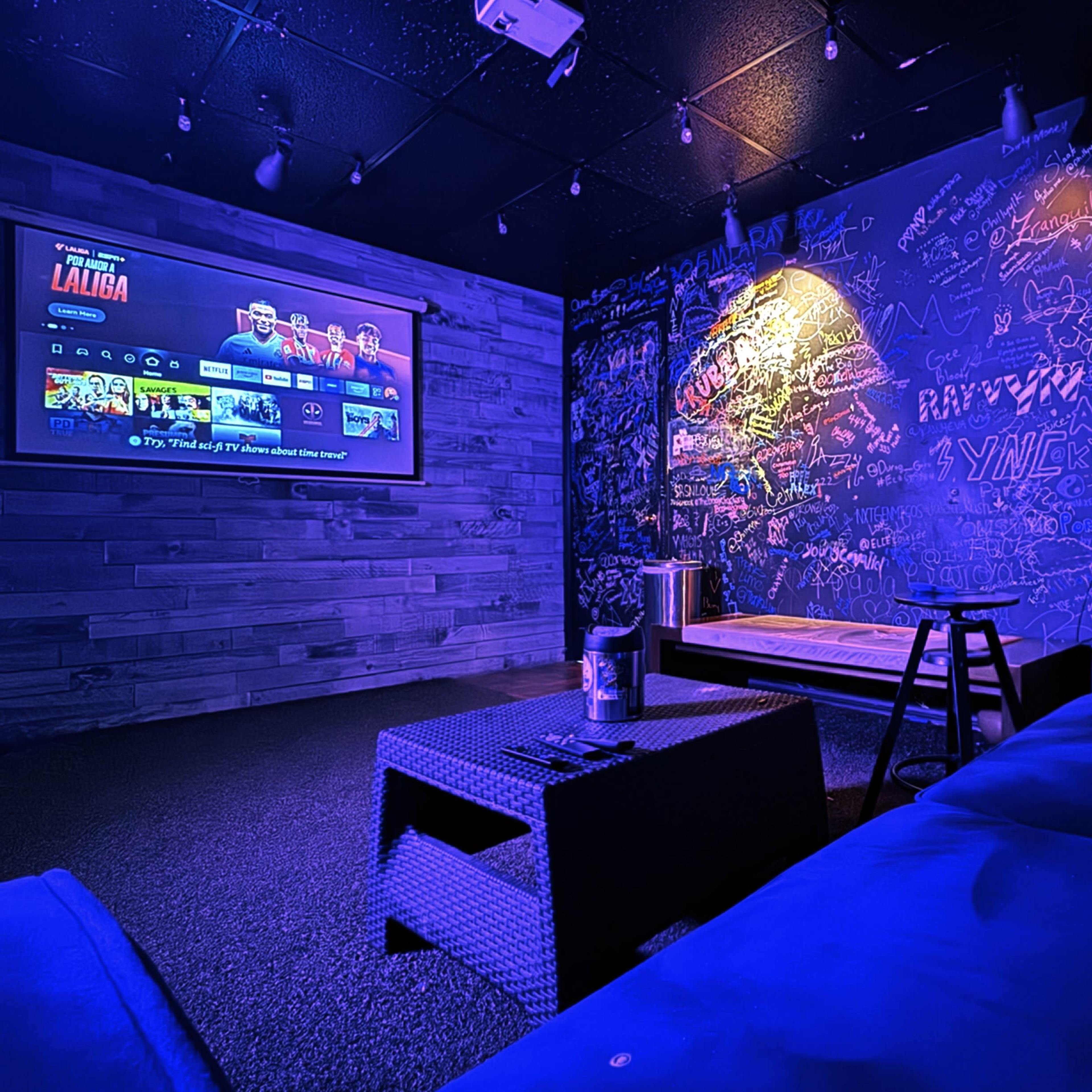 A cozy room with a large screen displaying entertainment options, a couch, a table, and walls covered in colorful graffiti.