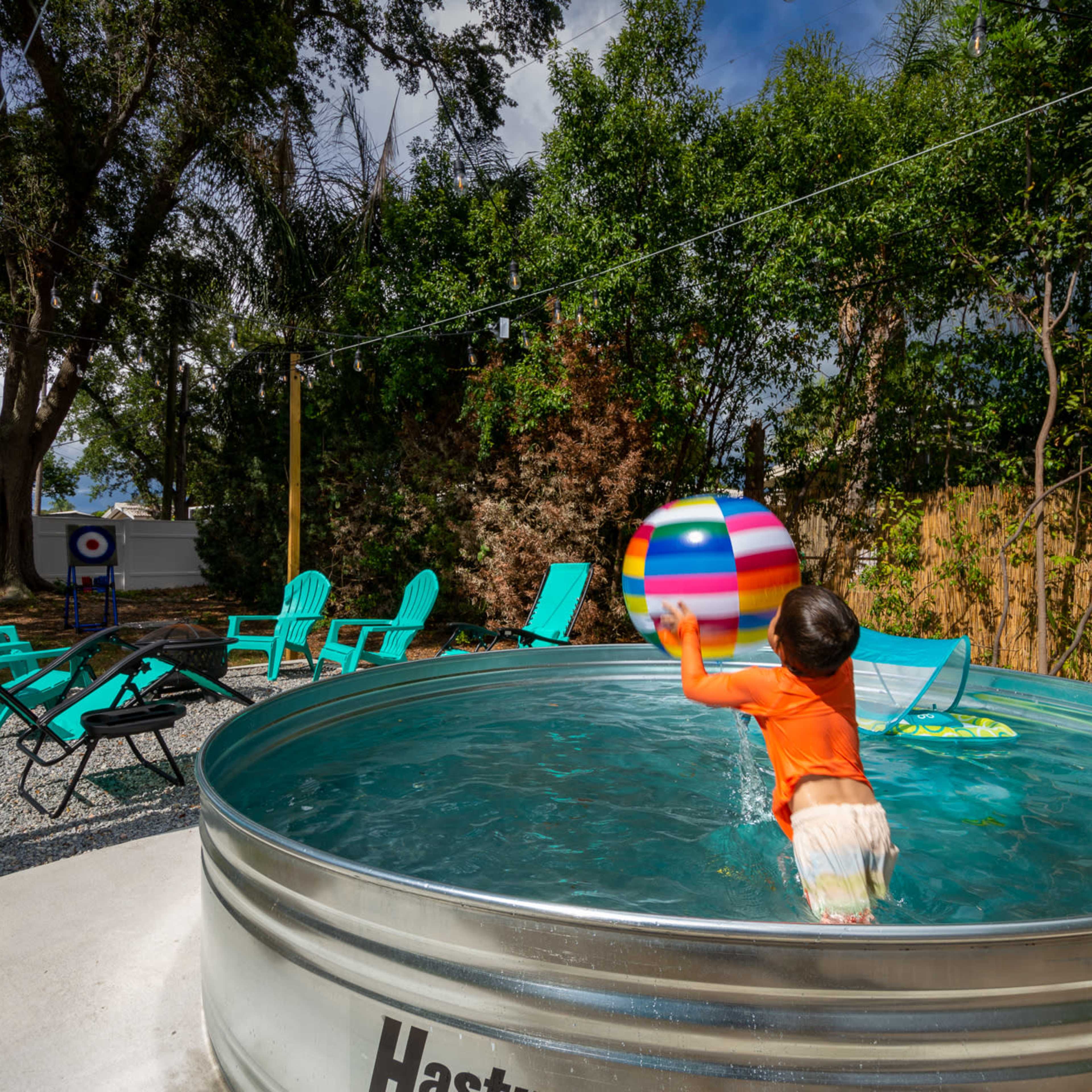 A child in an orange shirt plays with a colorful beach ball while standing in a metal pool surrounded by teal chairs and trees.