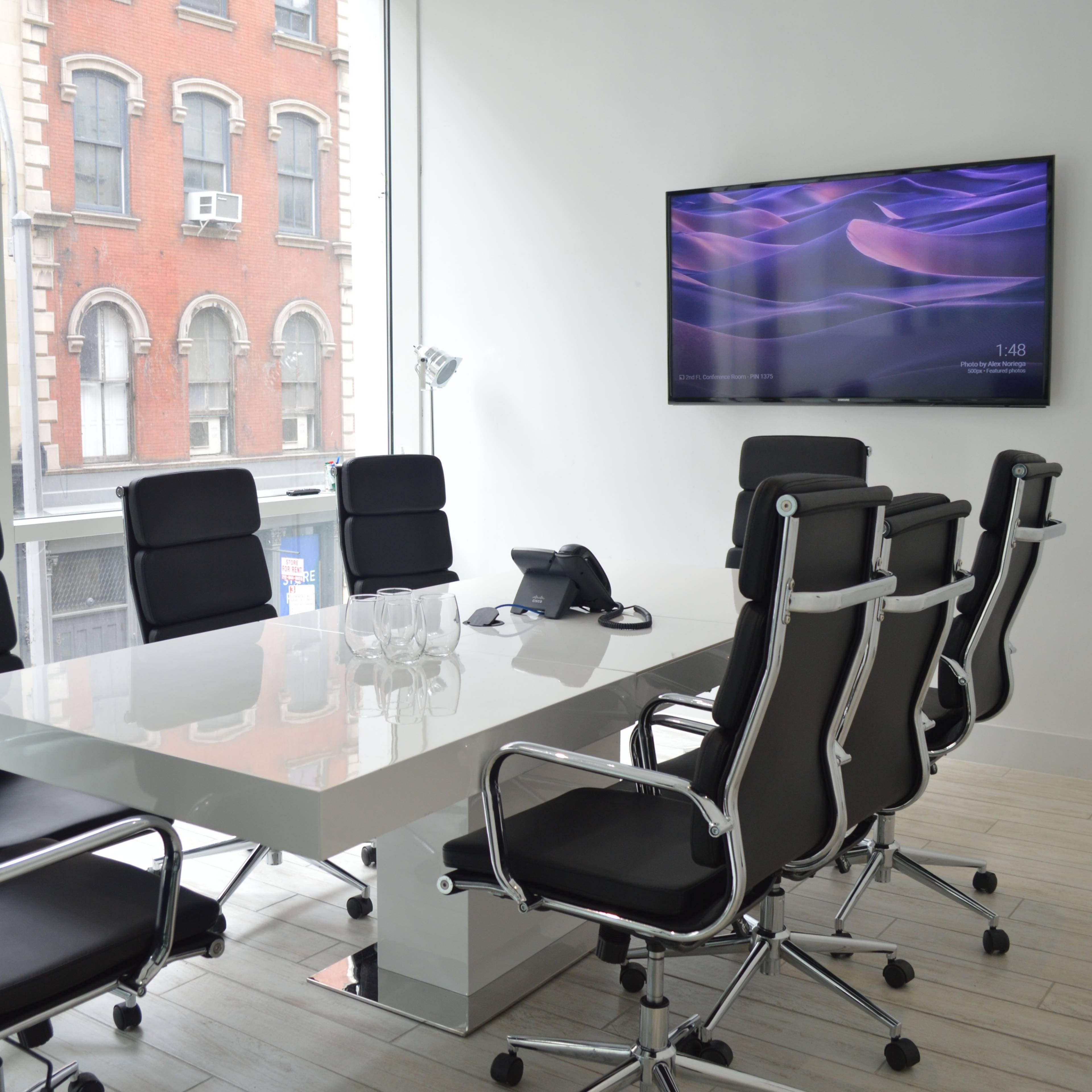 A modern conference room with a large table, eight black chairs, a telephone, and a wall-mounted television, overlooking a city street through a window.