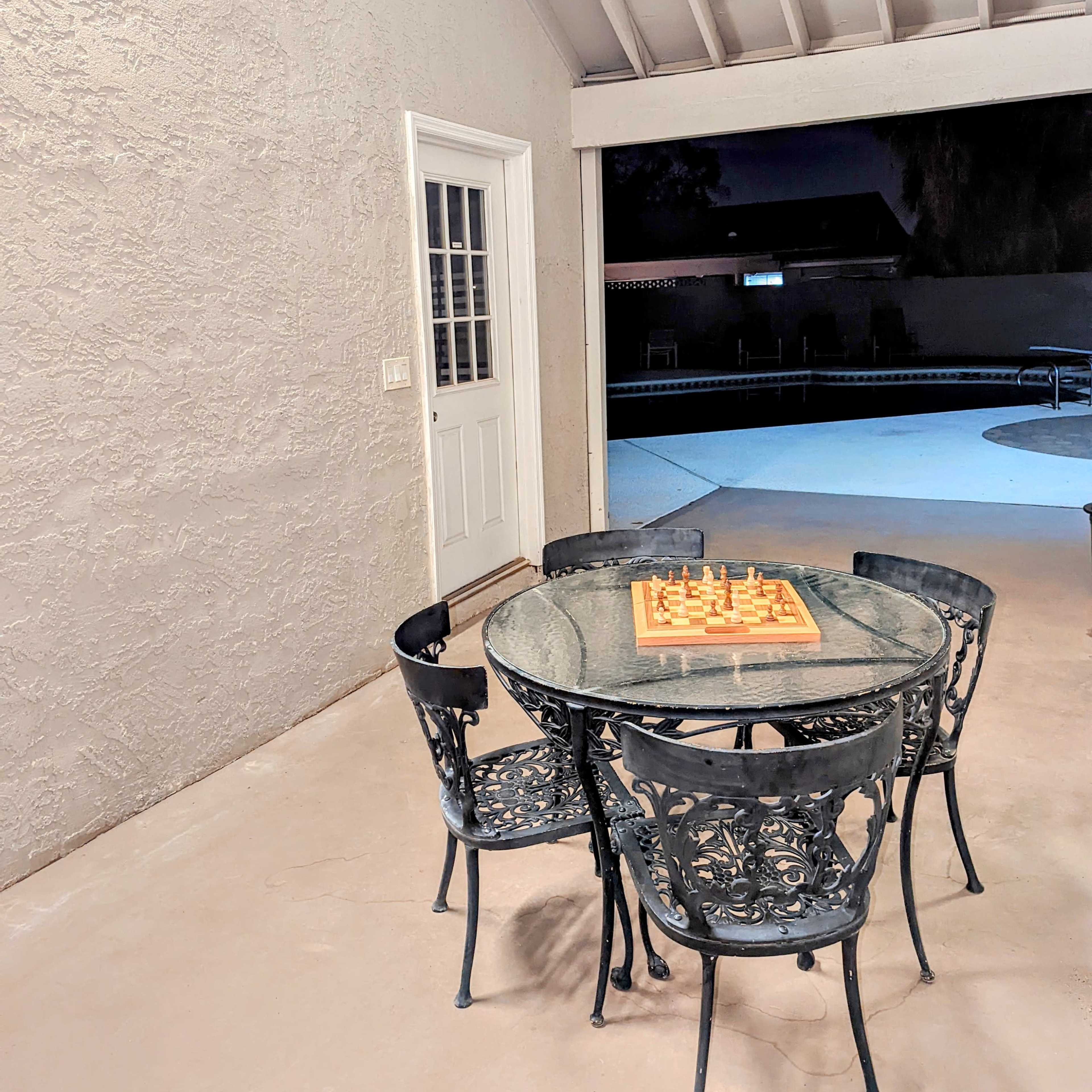 A round glass table with a chessboard sits surrounded by four black wrought iron chairs on a patio with a white ceiling.