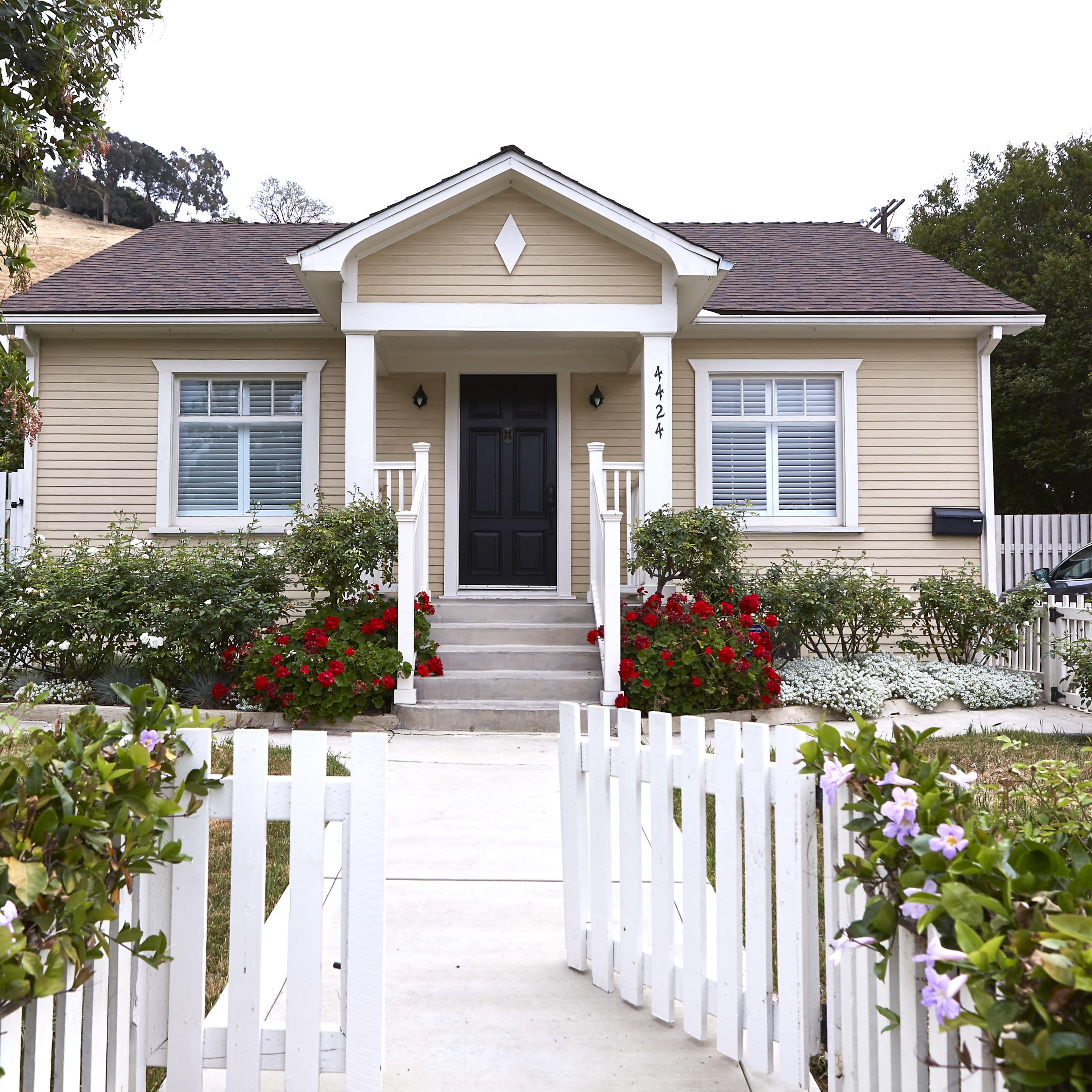 A small, light yellow house with a black front door is surrounded by a white picket fence and colorful flower beds.