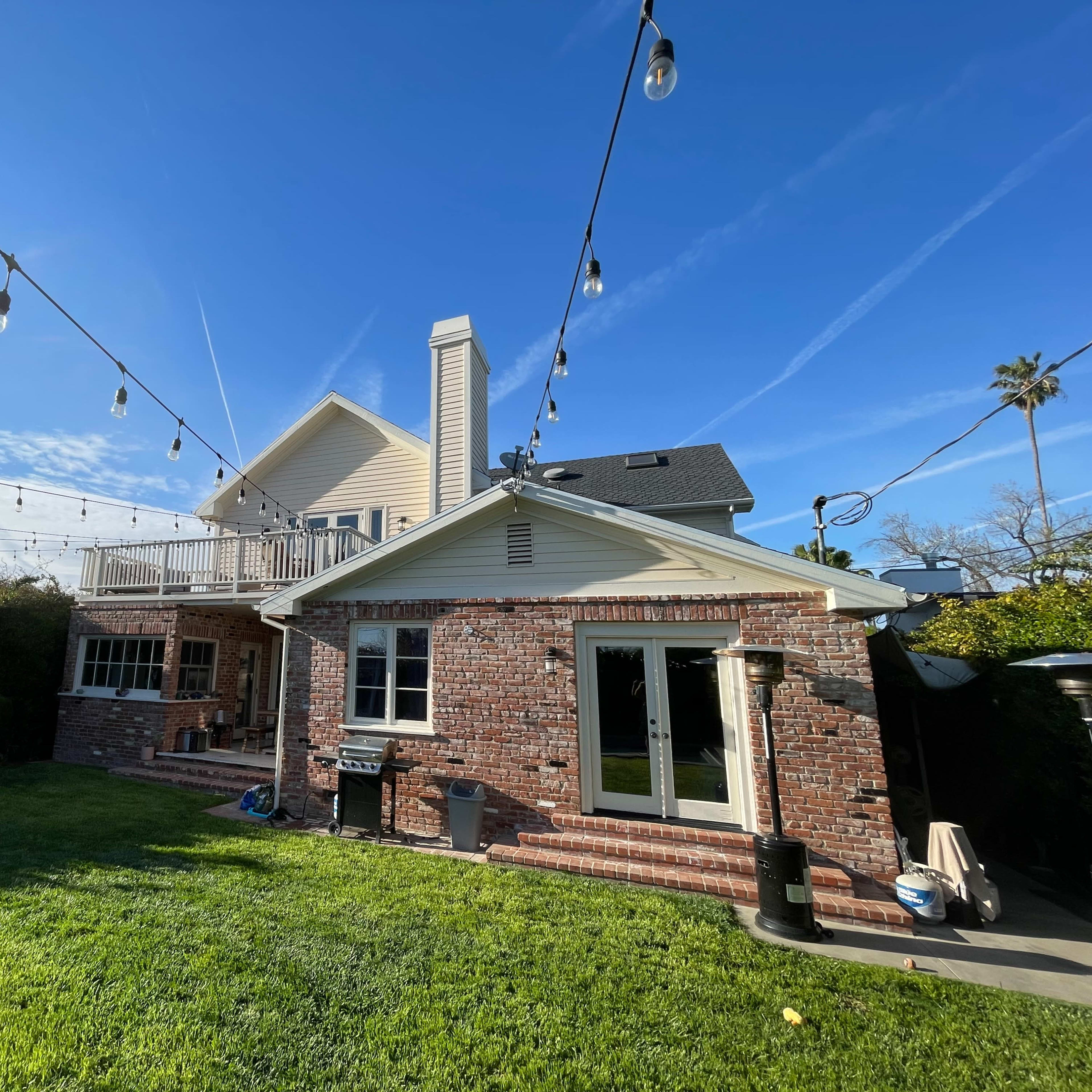 A two-story house with a brick wall and a large backyard, decorated with string lights overhead against a clear blue sky.