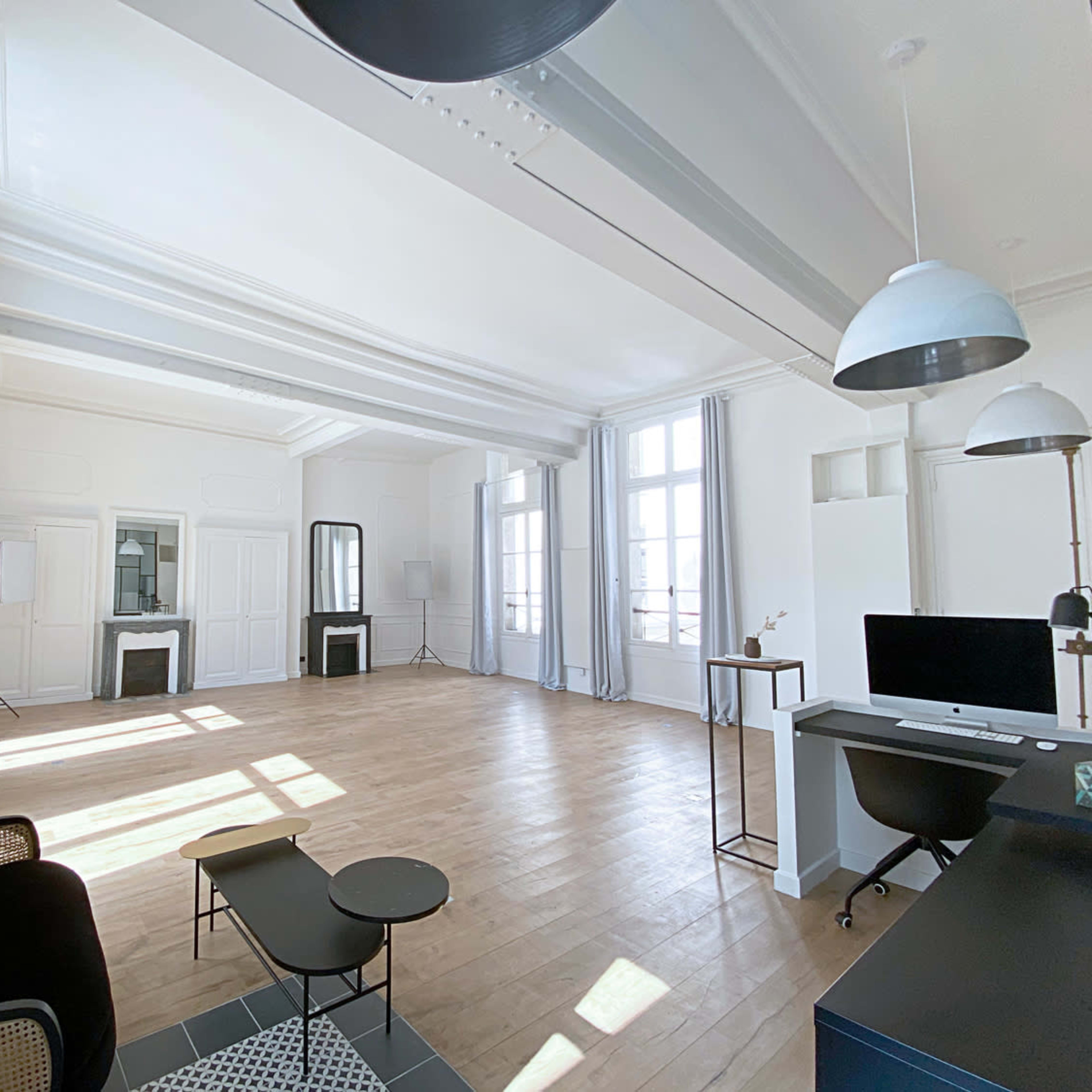 A spacious, sunlit office with light-colored walls, wooden flooring, and large windows, furnished with a black sofa, a glass coffee table, and a desk with a computer.