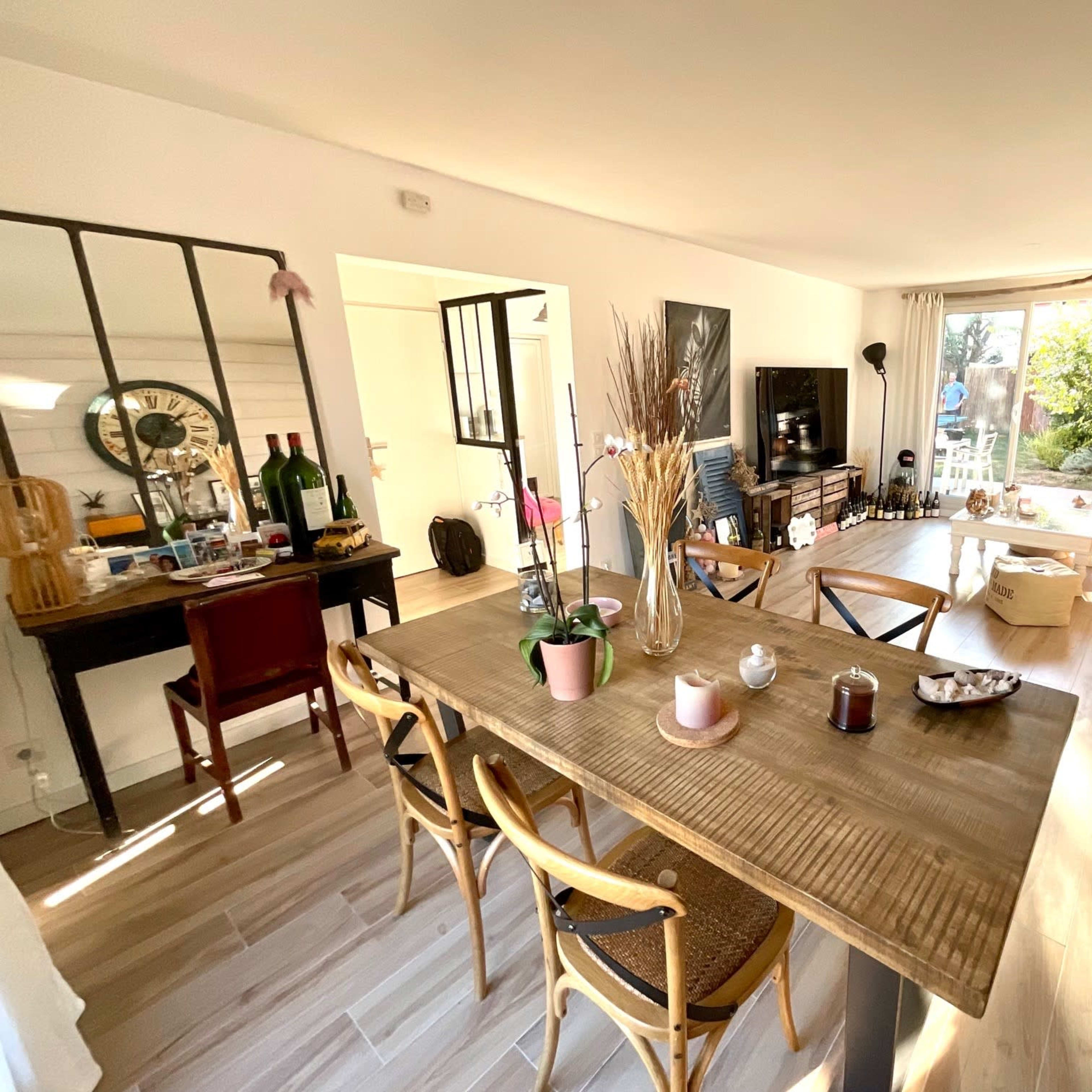 A bright living space features a wooden dining table surrounded by chairs, a desk with decor and bottles, and a cozy seating area with a fireplace and large windows.