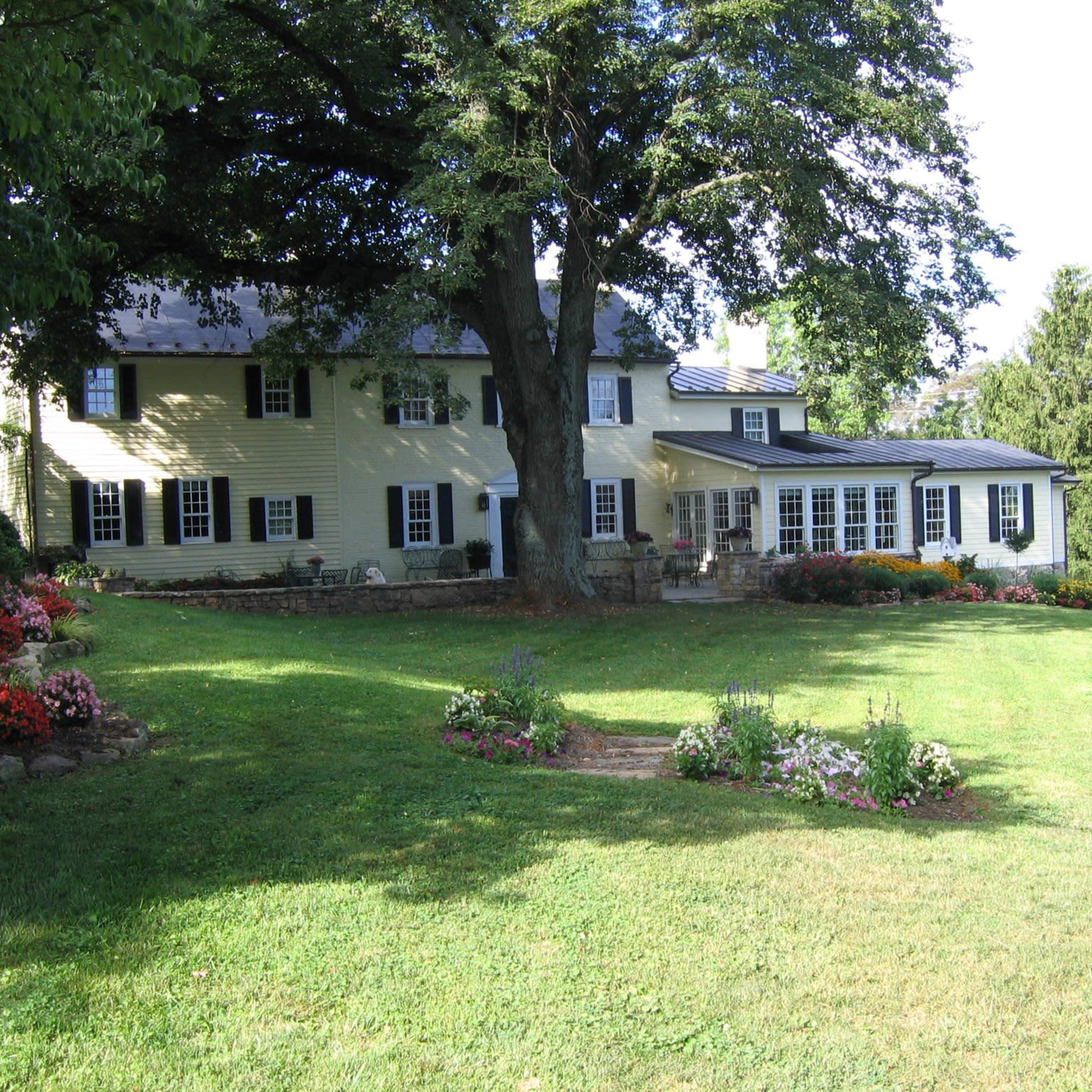 A two-story yellow house with a dark roof is surrounded by a landscaped yard featuring flower beds and large trees.
