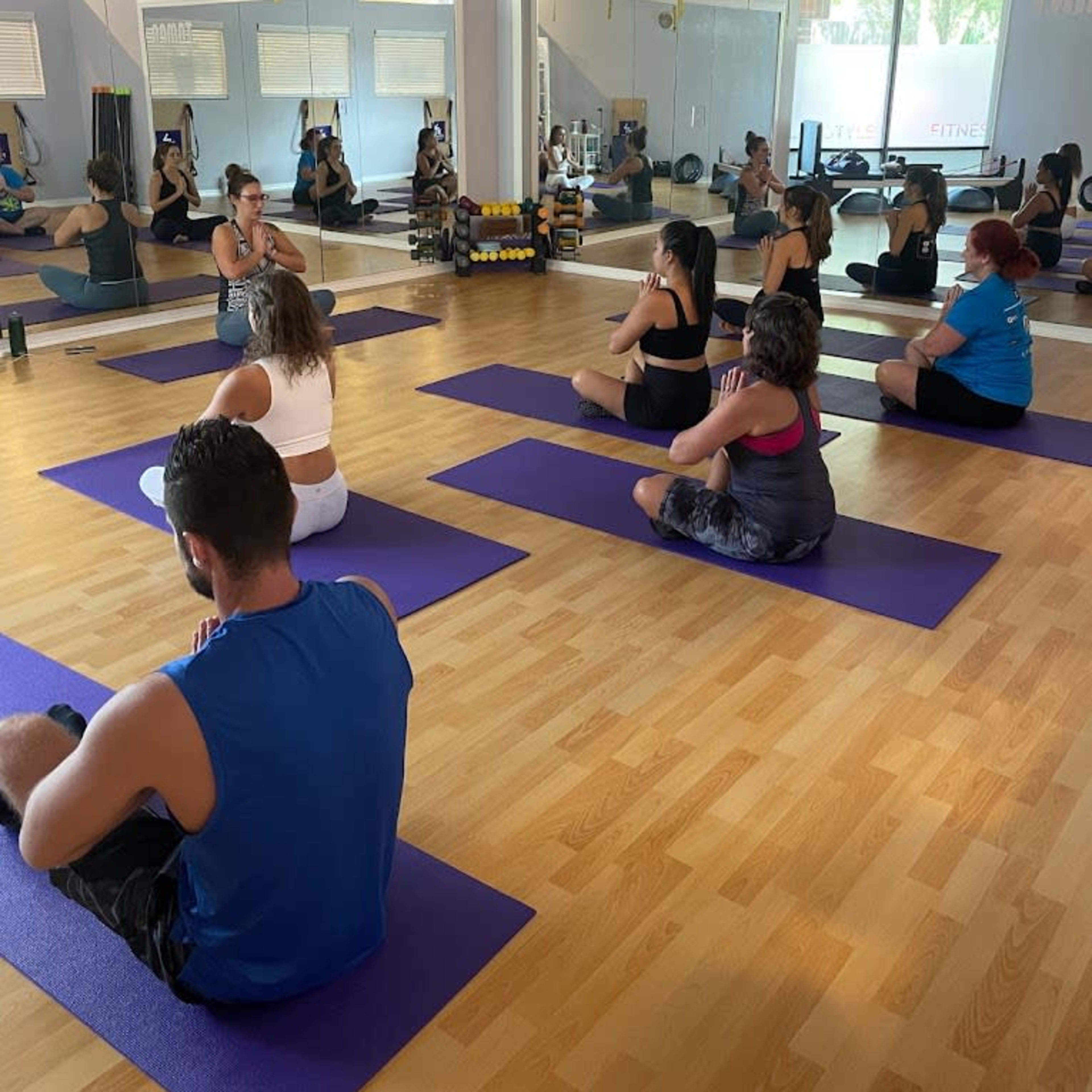 A group of people is practicing yoga on purple mats in a well-lit studio with mirrors and fitness equipment in the background.
