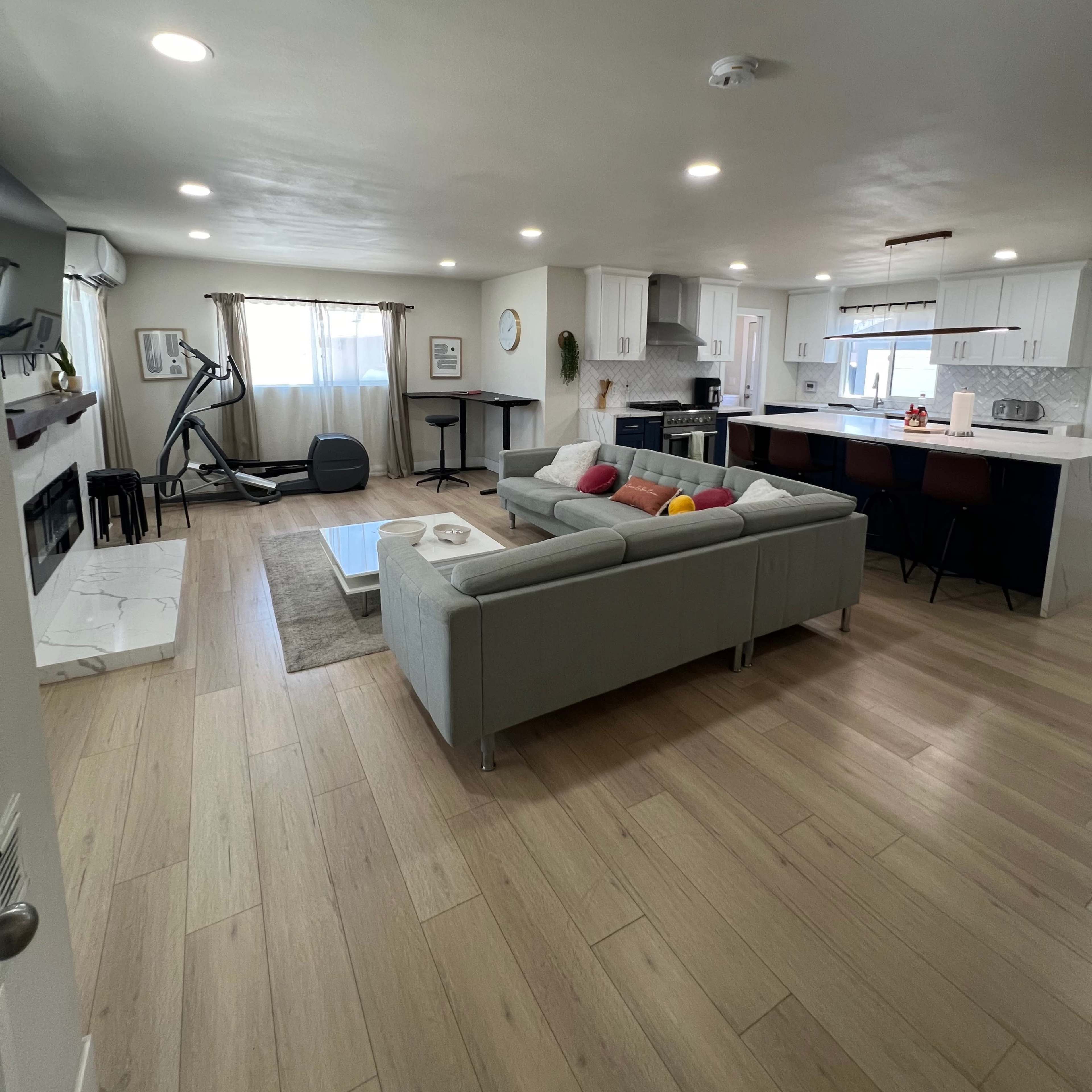 A modern living room with a sofa, a coffee table, a fitness machine, and a kitchen area in the background.