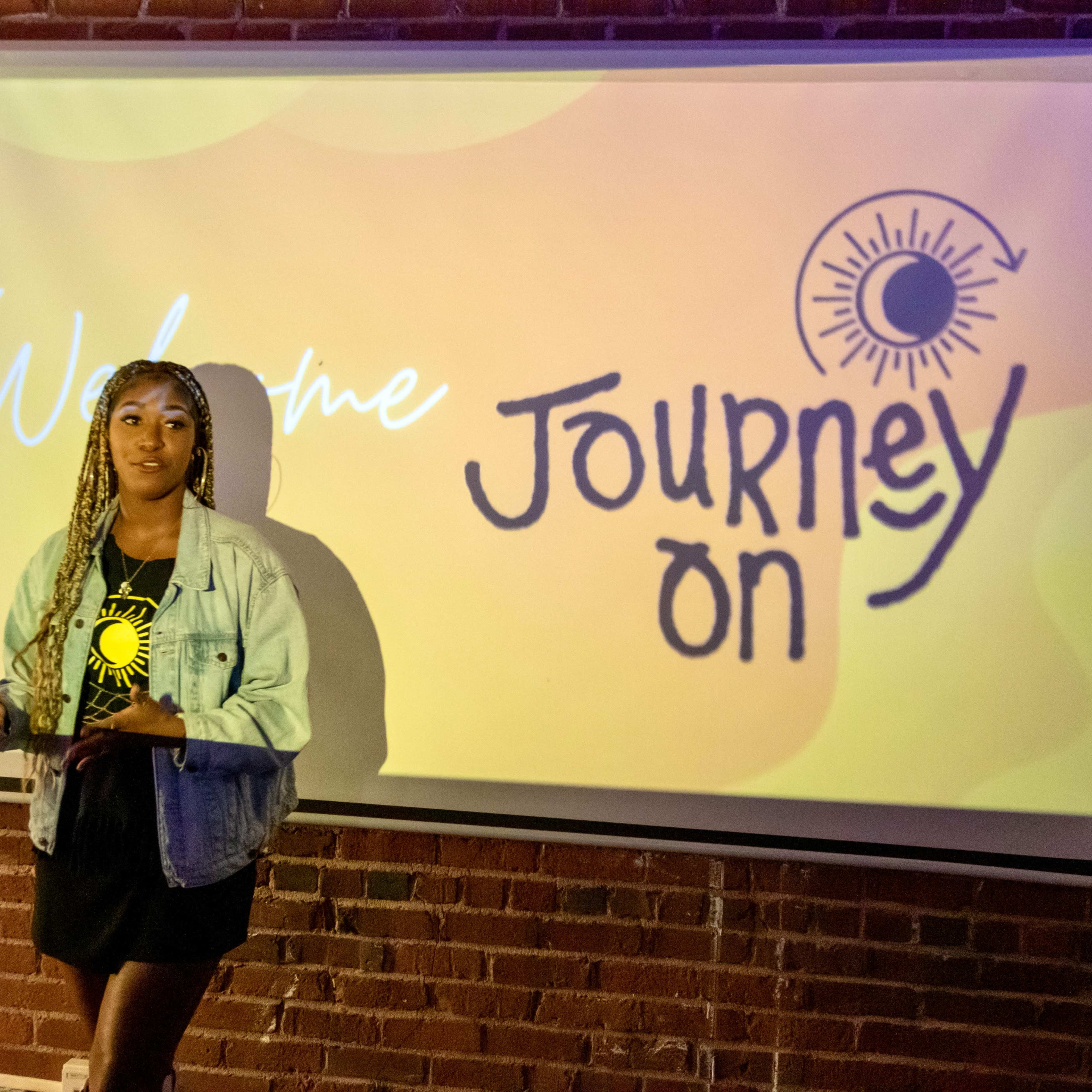A woman stands in front of a projector screen displaying the words "Welcome" and "Journey On" against a vibrant background in a brick-walled room.