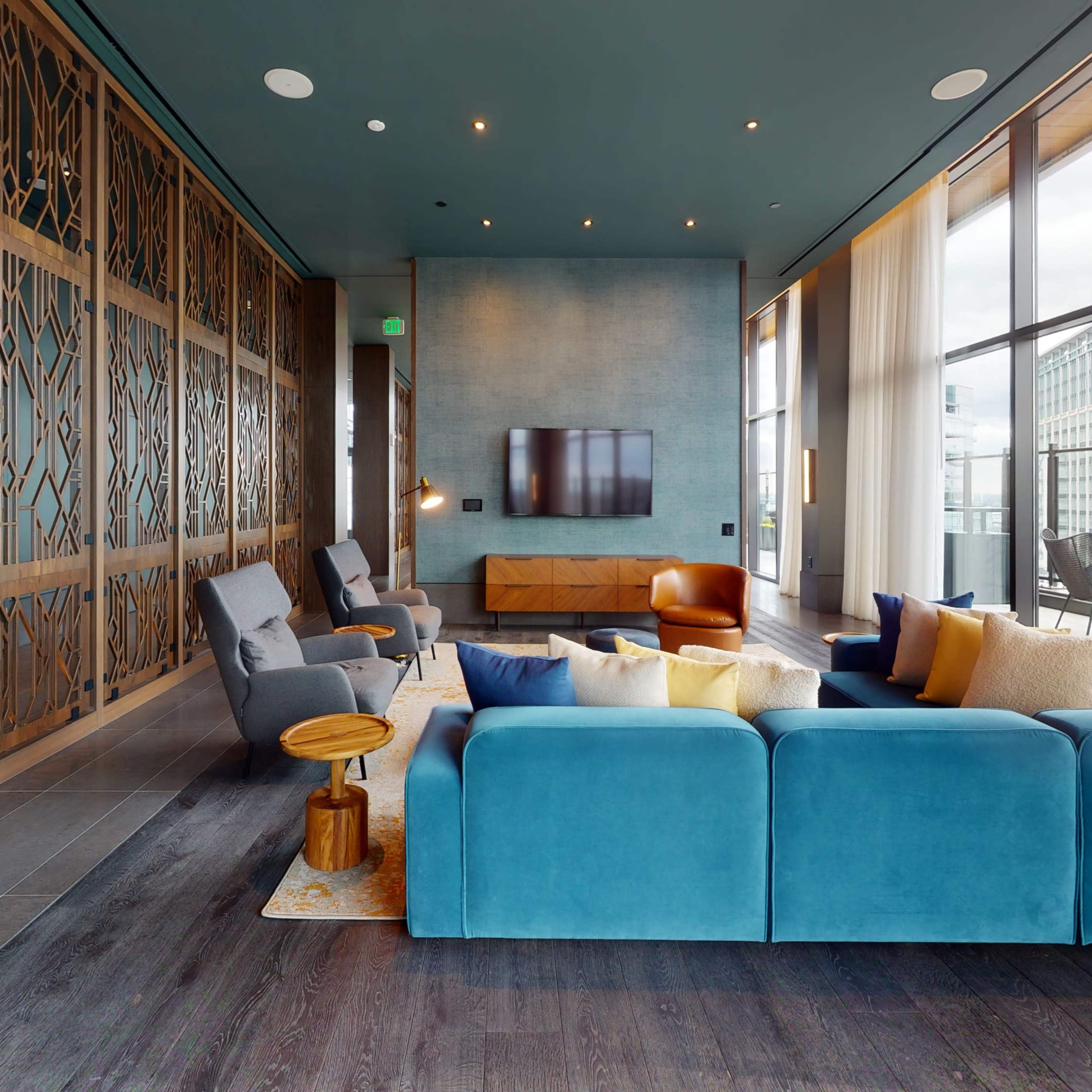 A modern lounge area with large windows, a wall-mounted TV, and a mix of blue and gray furniture accented with decorative pillows.