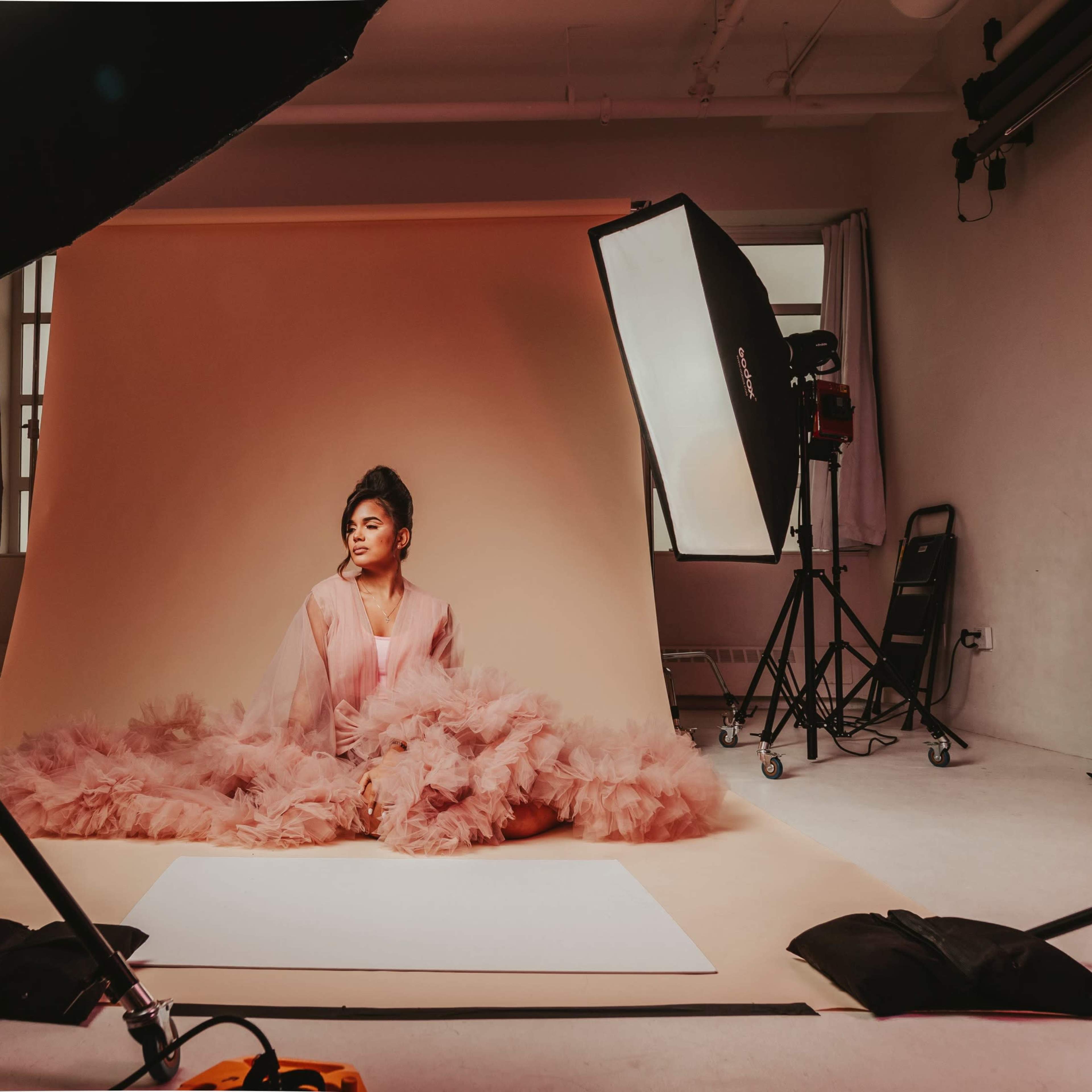 A model in a pink outfit sits on the floor in a photography studio with soft lighting and a beige backdrop.