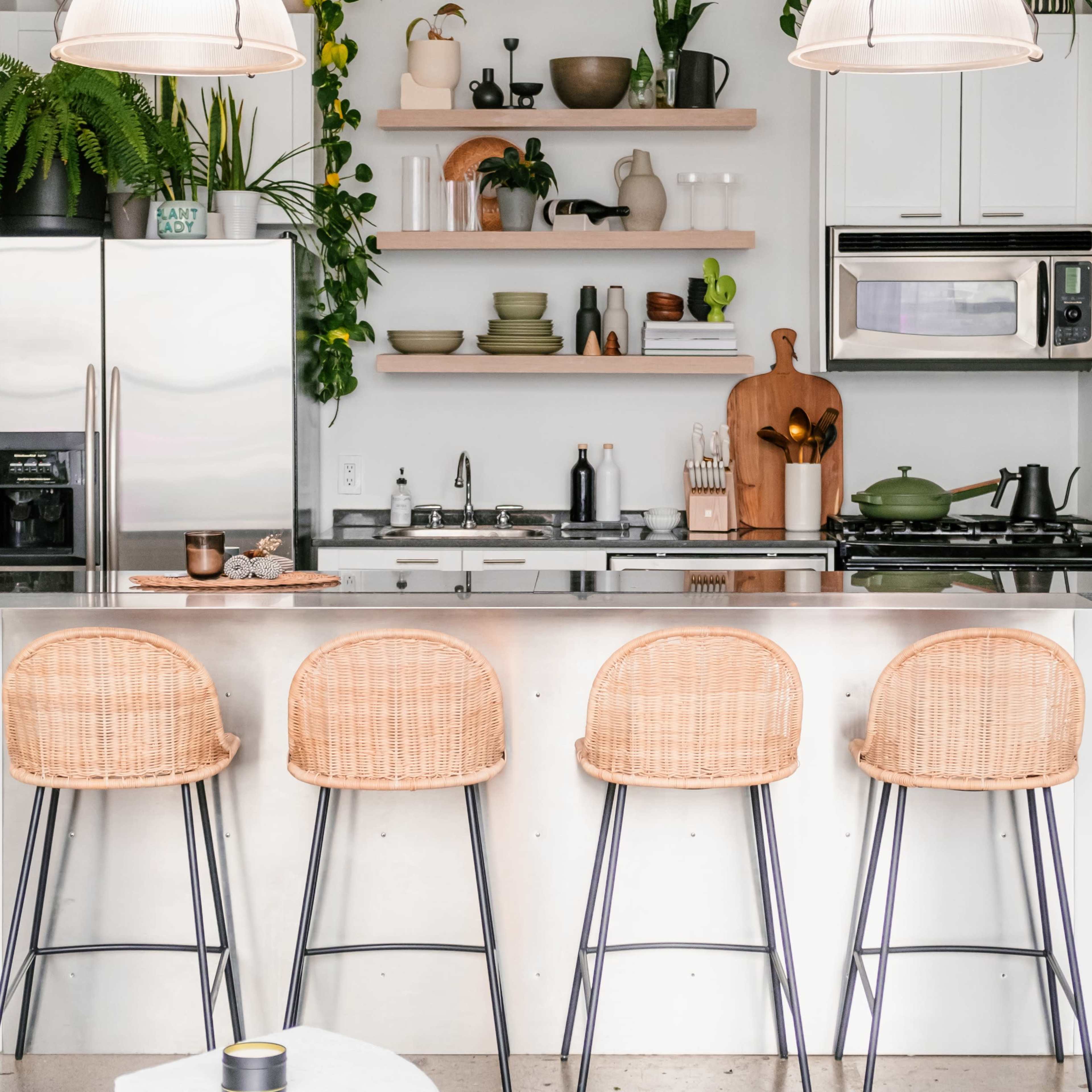 A modern kitchen features a sleek island bar with four woven stools, surrounded by plants and organized shelves displaying kitchenware and decor.