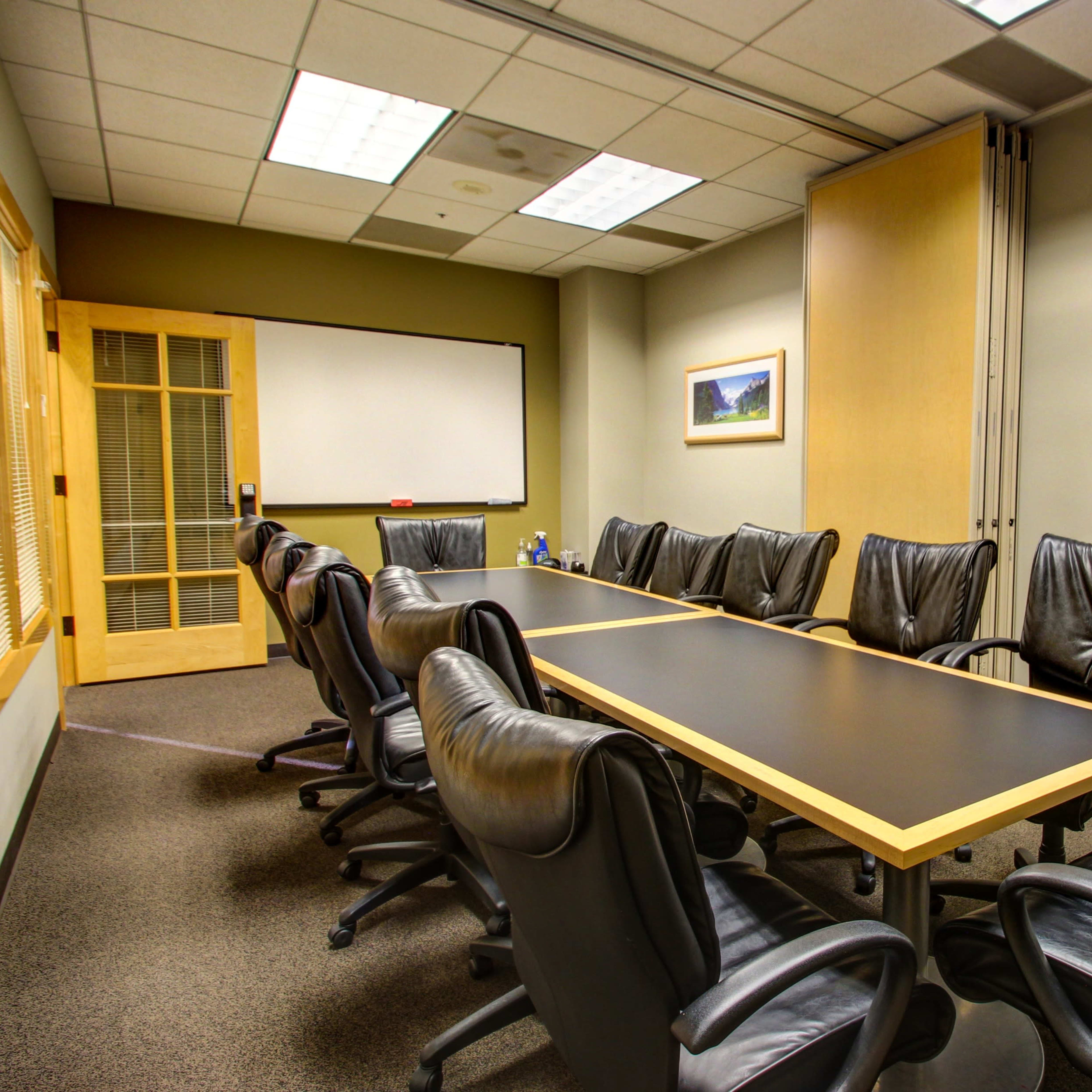 A conference room is furnished with a long table surrounded by black leather chairs, featuring a whiteboard and framed landscape pictures on the walls.
