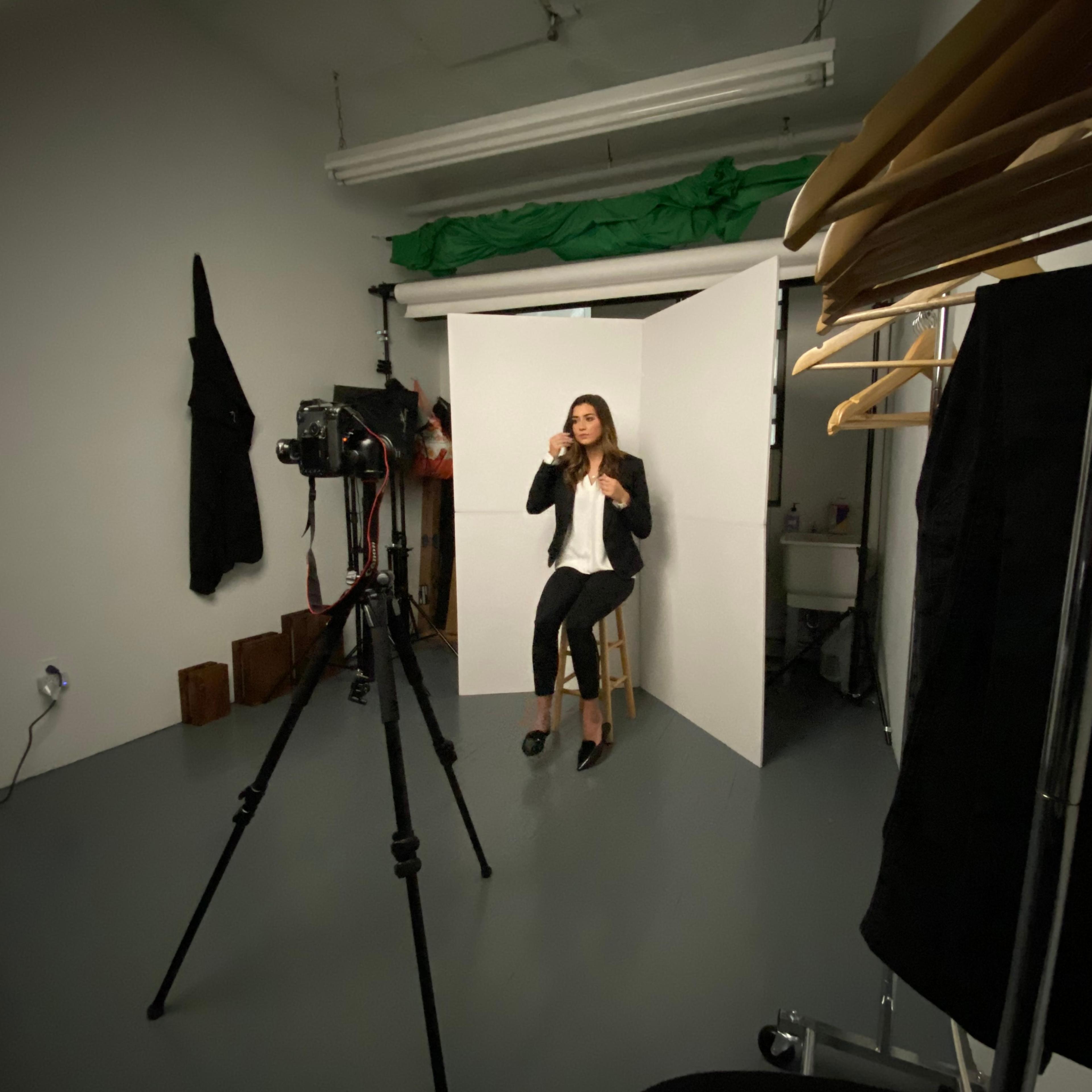 A person sits on a stool in a photo studio, posing in front of a camera set up on a tripod, with a backdrop and clothing racks visible in the scene.