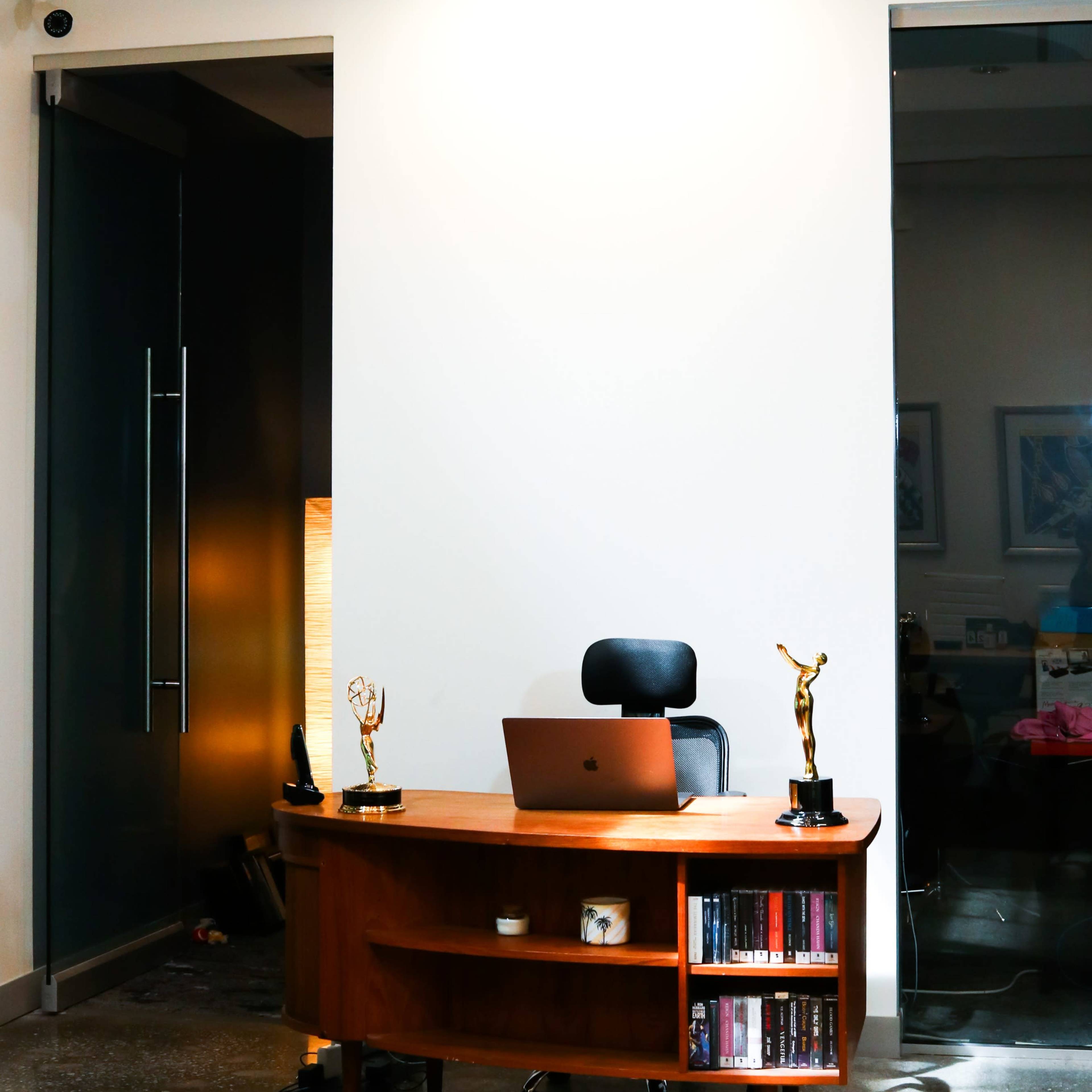 A modern workspace features a wooden desk with a laptop and two trophy statuettes, positioned near a large glass entryway and an elaborate ceiling light fixture.