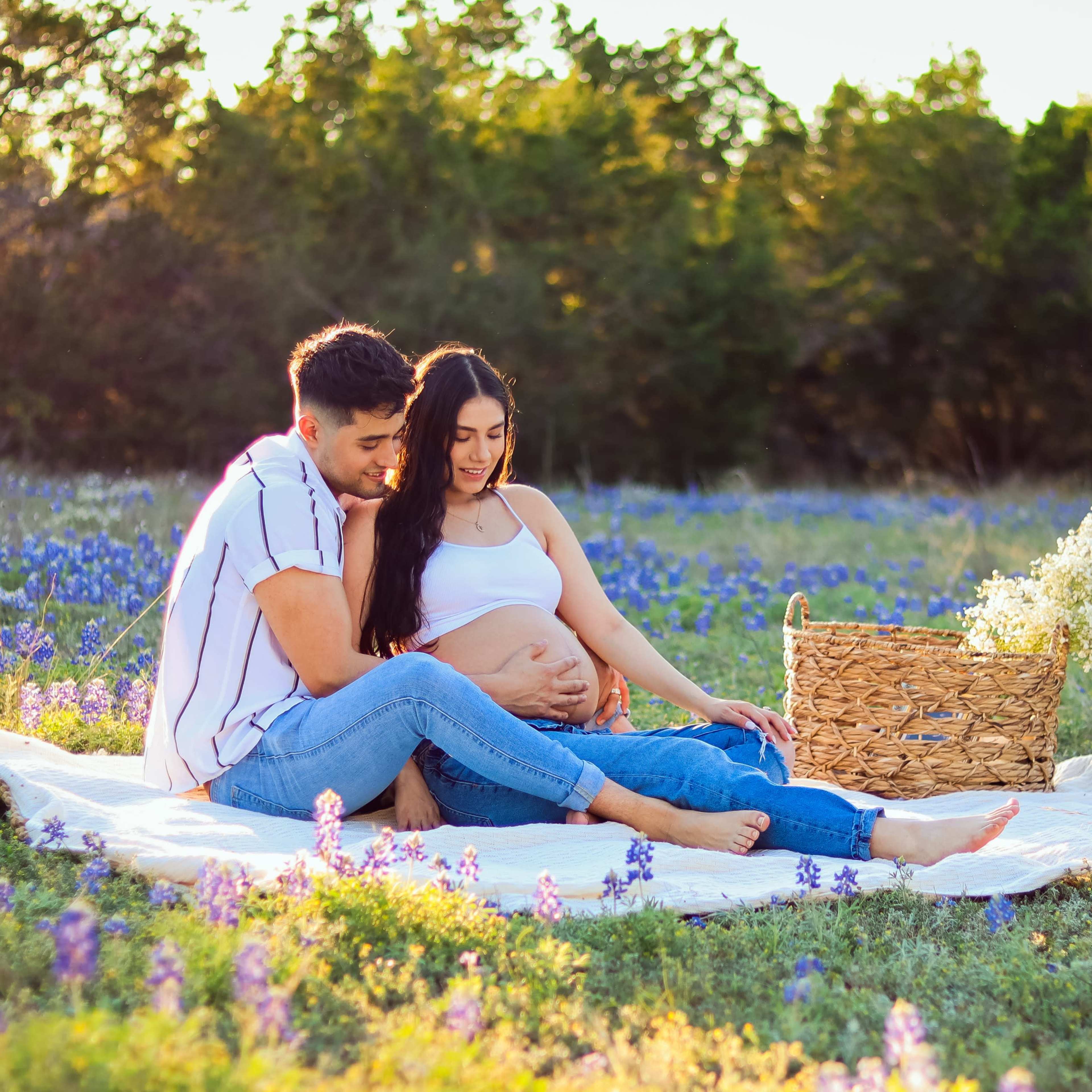 A couple sits on a blanket in a field of bluebonnets, with the woman cradling her pregnant belly and a basket of flowers beside them.