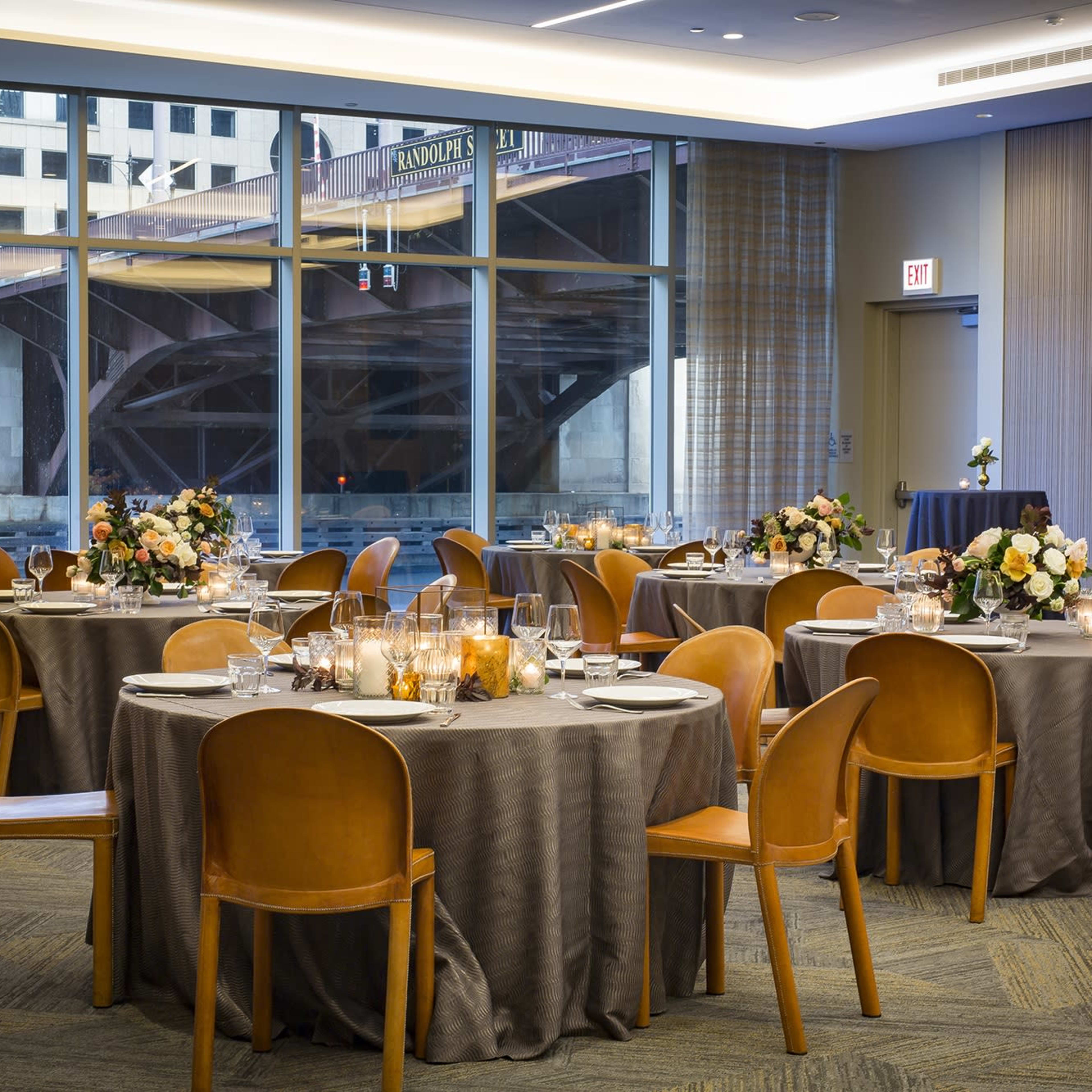 A dining room with several round tables set for a meal, featuring tablecloths, flower arrangements, and a view of a bridge through large windows.