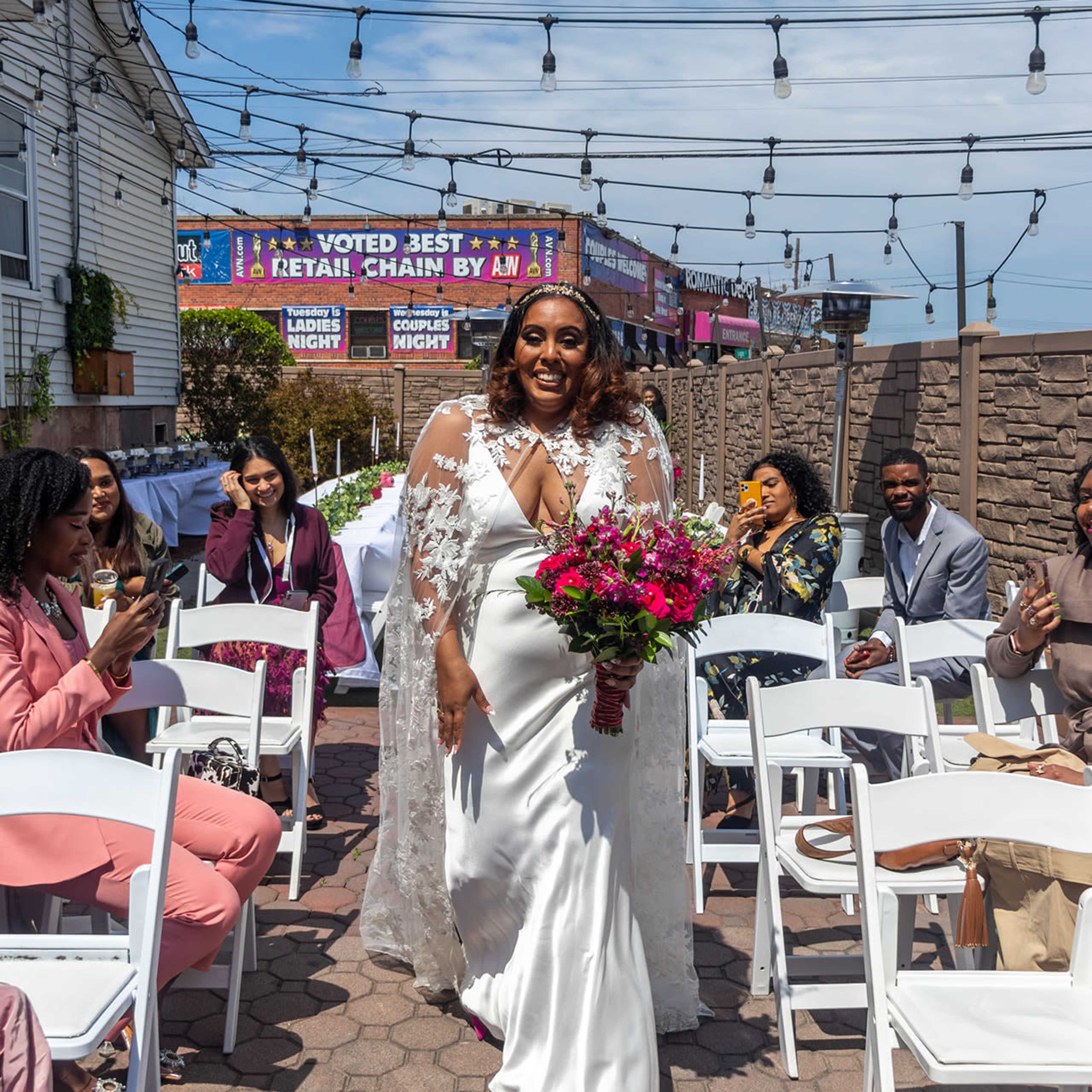 A bride in a white gown walks down an aisle surrounded by seated guests, holding a bouquet of flowers amidst a decorated outdoor setting.