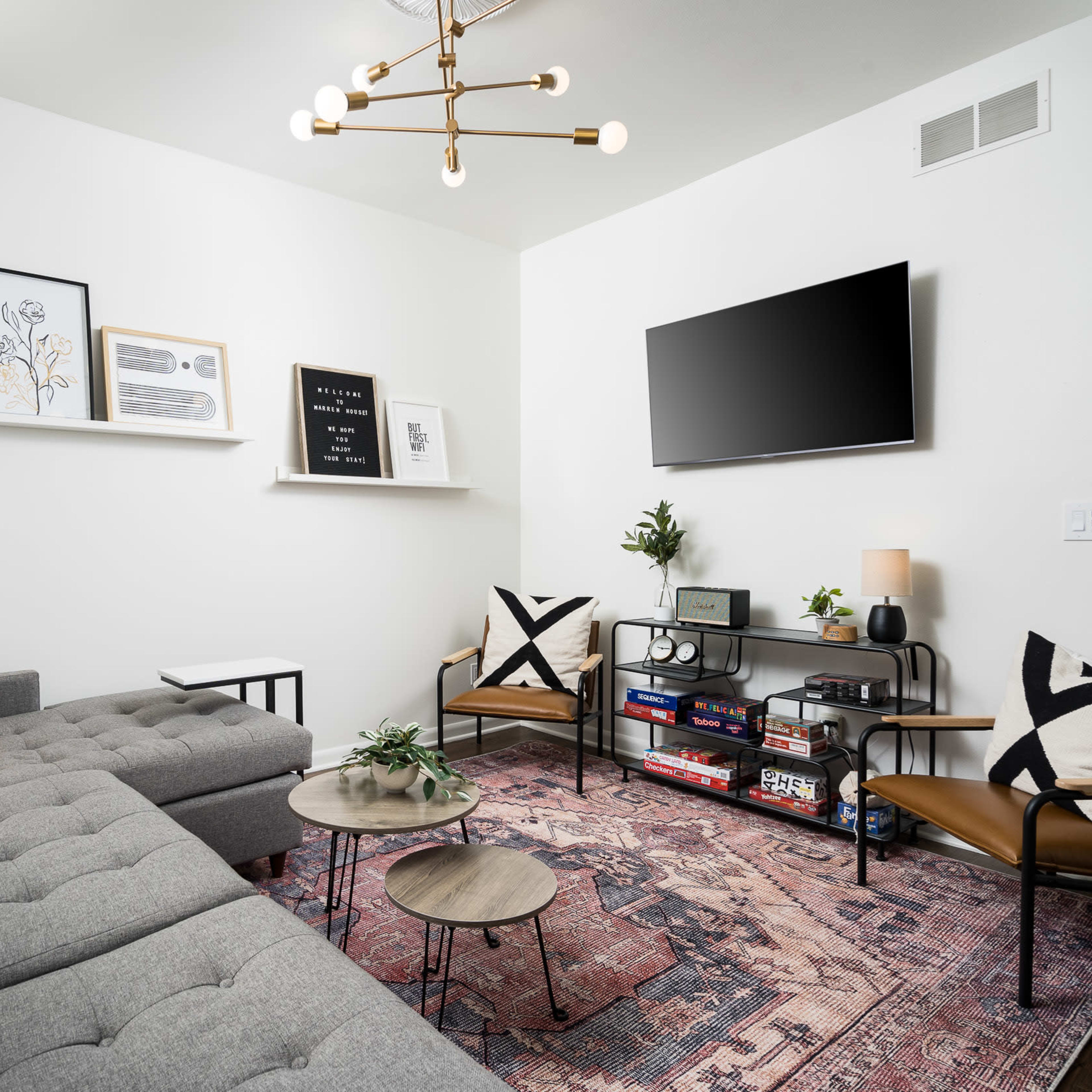 A cozy living room features a gray sectional sofa, mid-century chairs, a patterned rug, and various decorative elements on the walls and shelves.