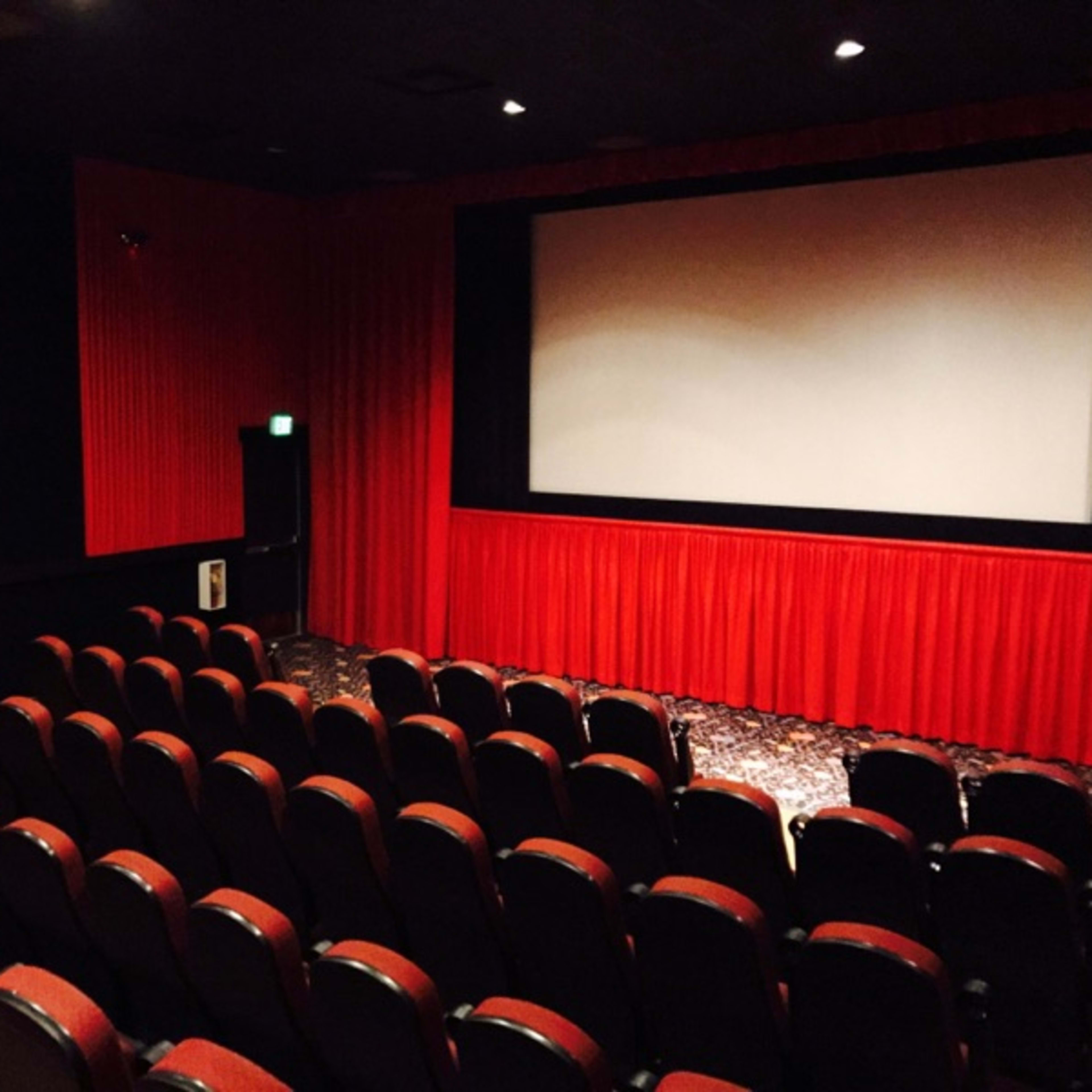 An empty movie theater with red curtains and rows of black seats facing a blank screen.