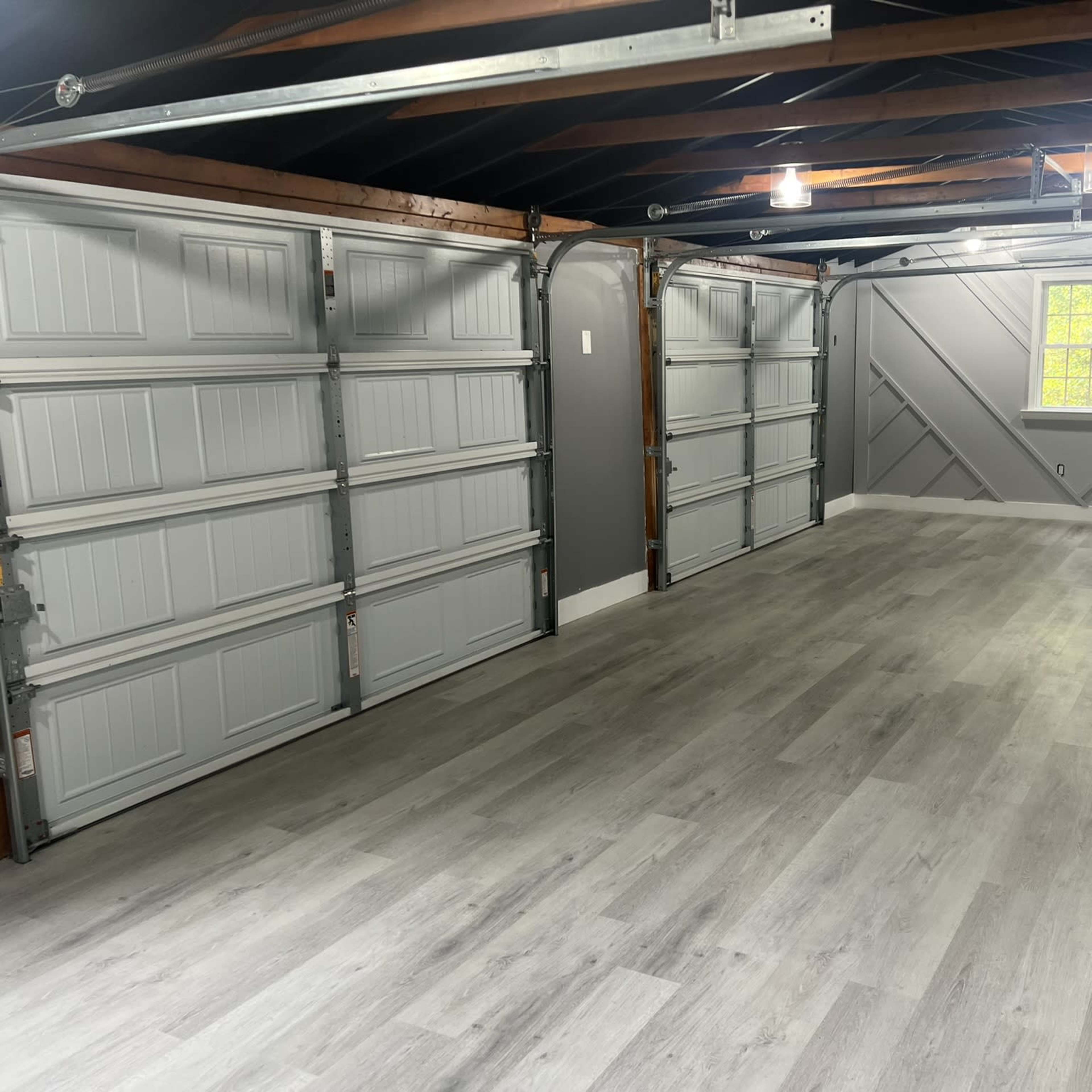 An empty garage with two closed garage doors and a window, featuring gray walls and a light-colored wood floor.