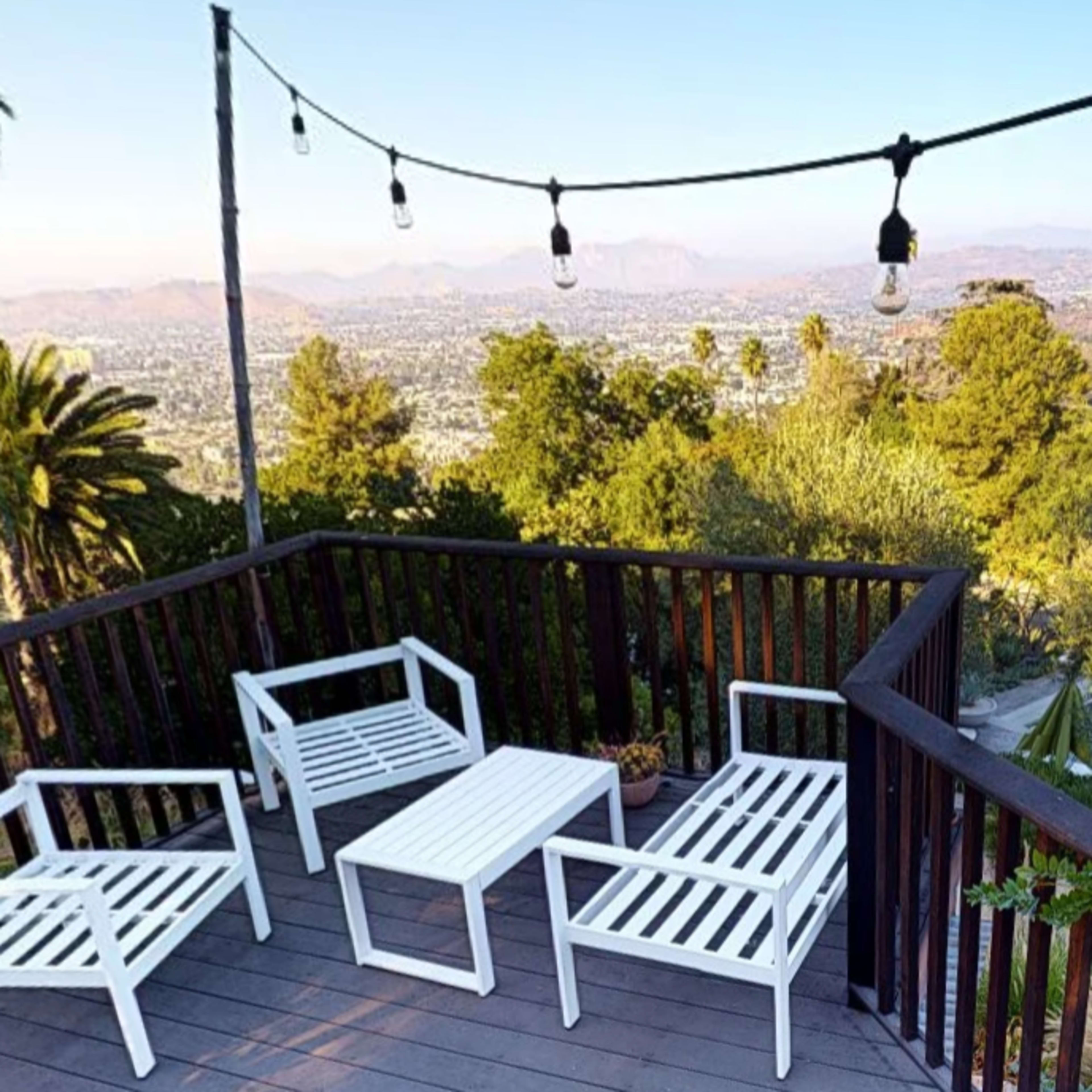 A deck with white patio furniture overlooks a valley surrounded by trees and mountains, with string lights overhead.