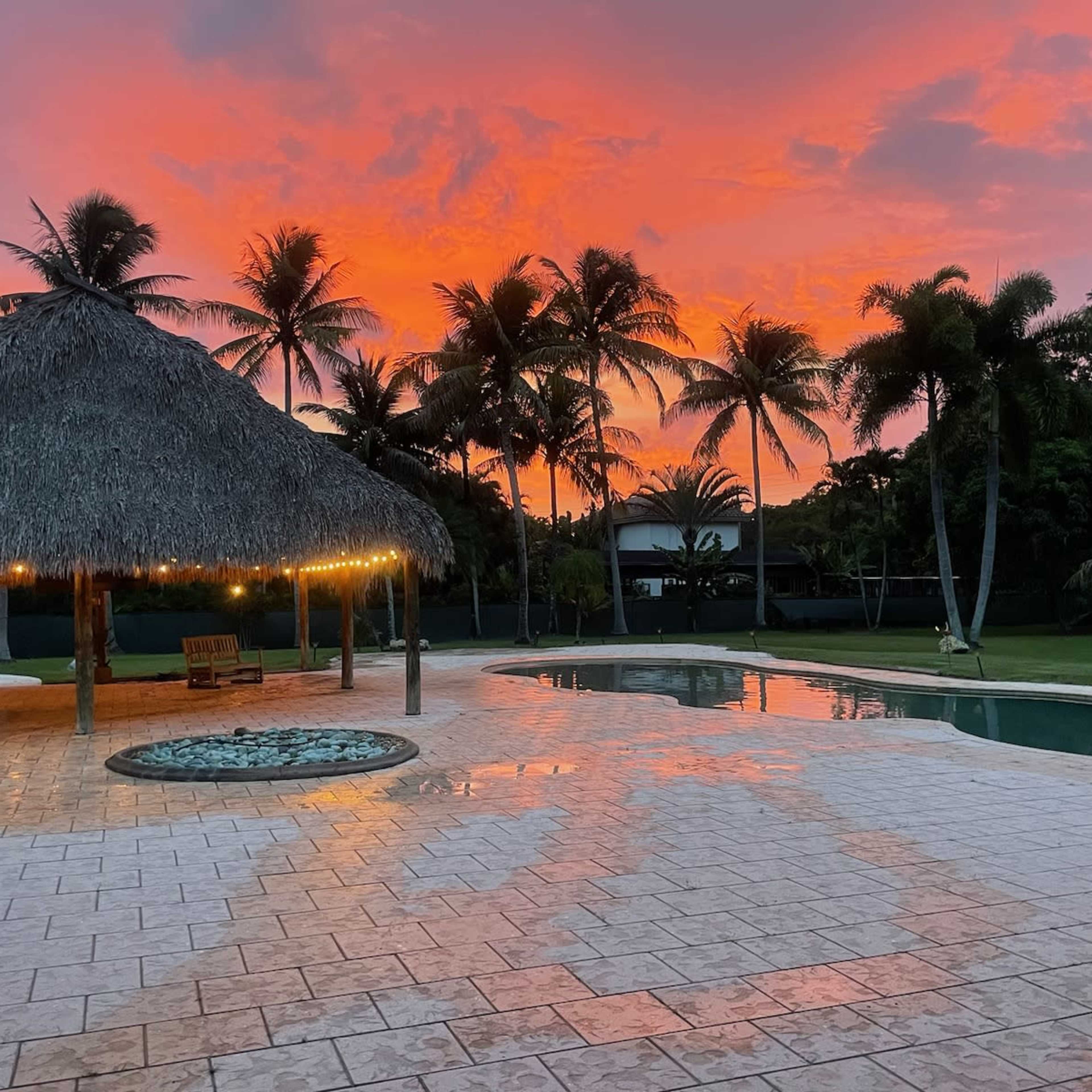 The scene depicts a pool area with a thatched gazebo surrounded by palm trees, reflecting the vibrant colors of a sunset.