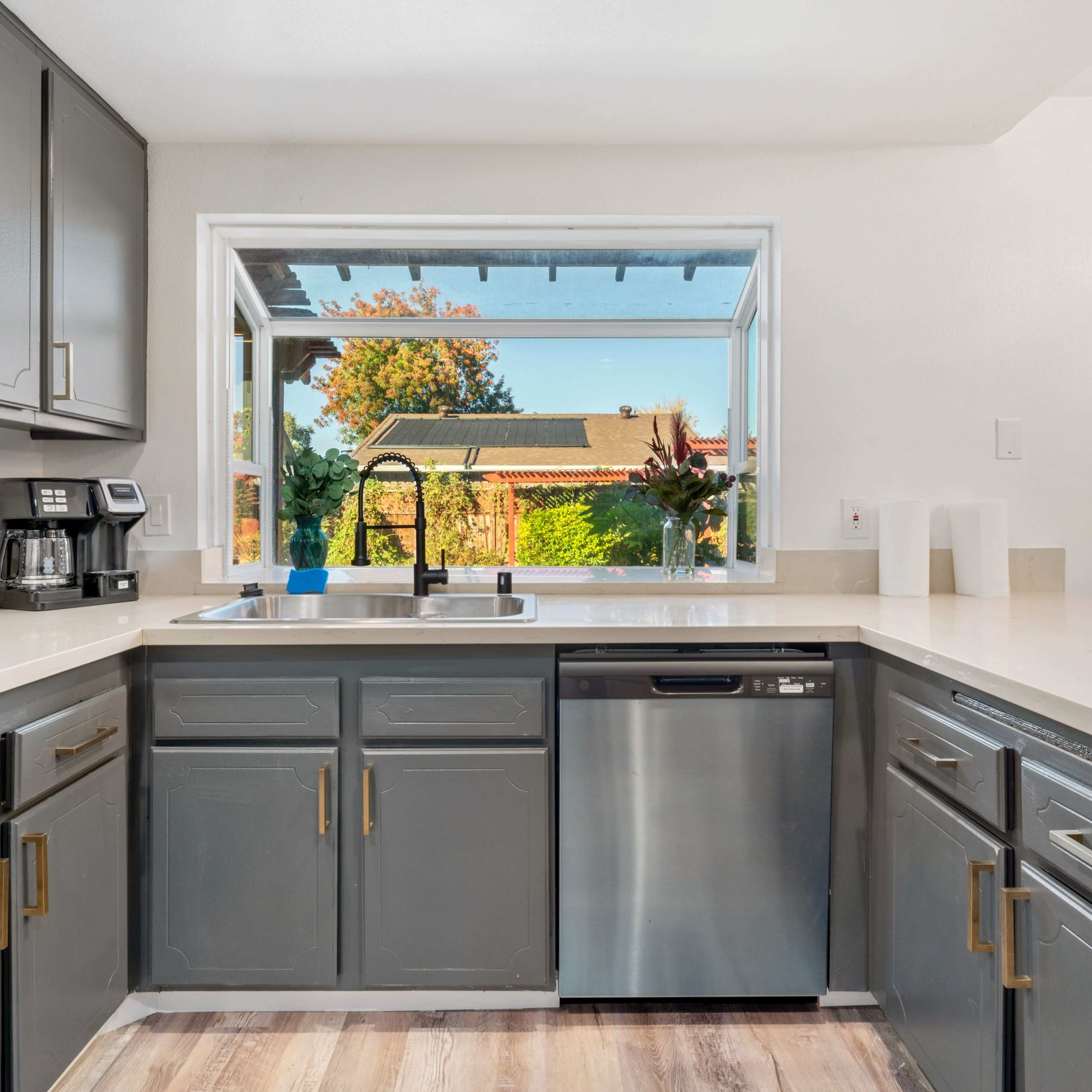A modern kitchen features gray cabinets, stainless steel appliances, and a large window overlooking a garden.