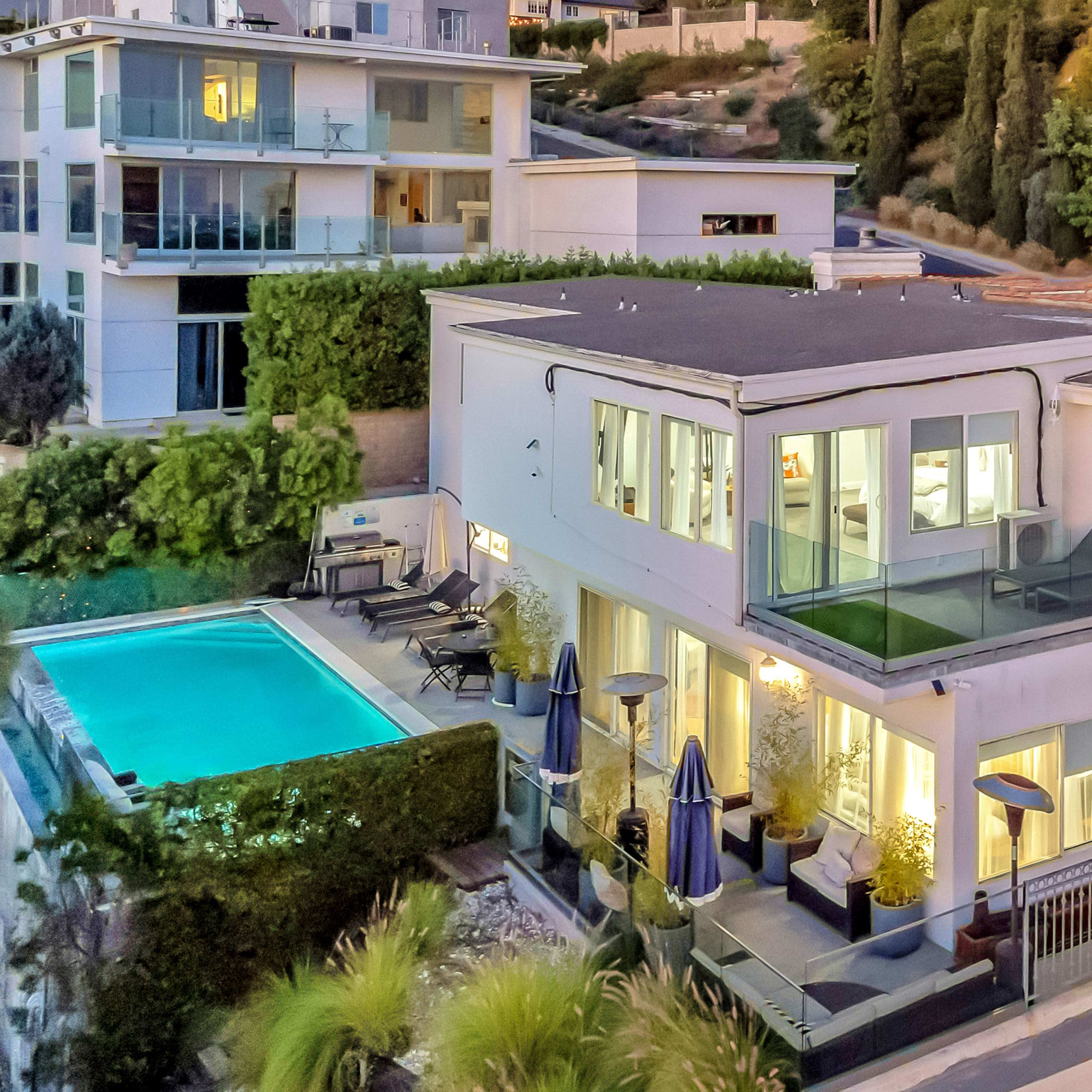 A modern house with large windows overlooks a pool and landscaped yard, situated amidst hilly terrain.