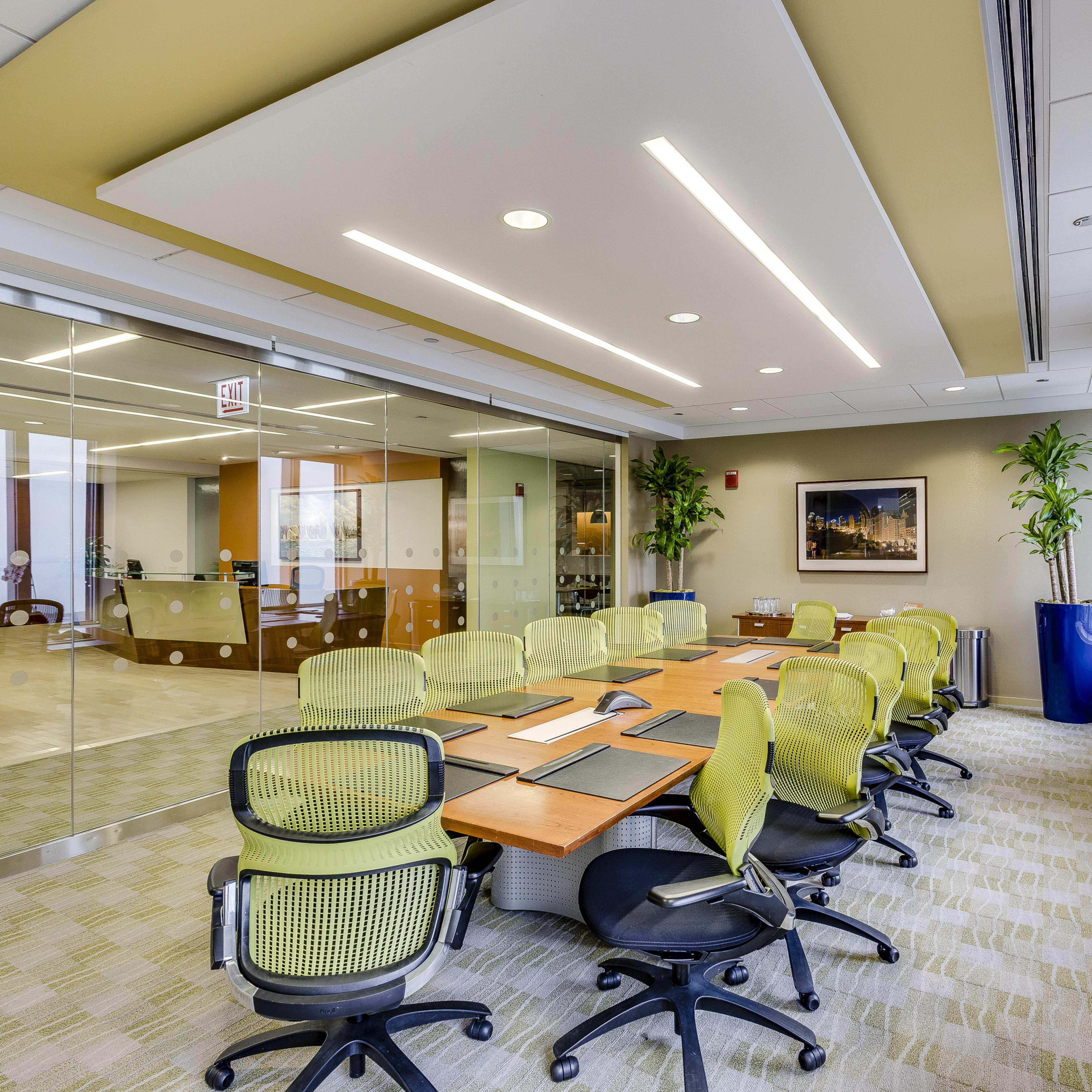 A modern conference room features a long wooden table surrounded by green mesh chairs, with large windows and plants enhancing the space.