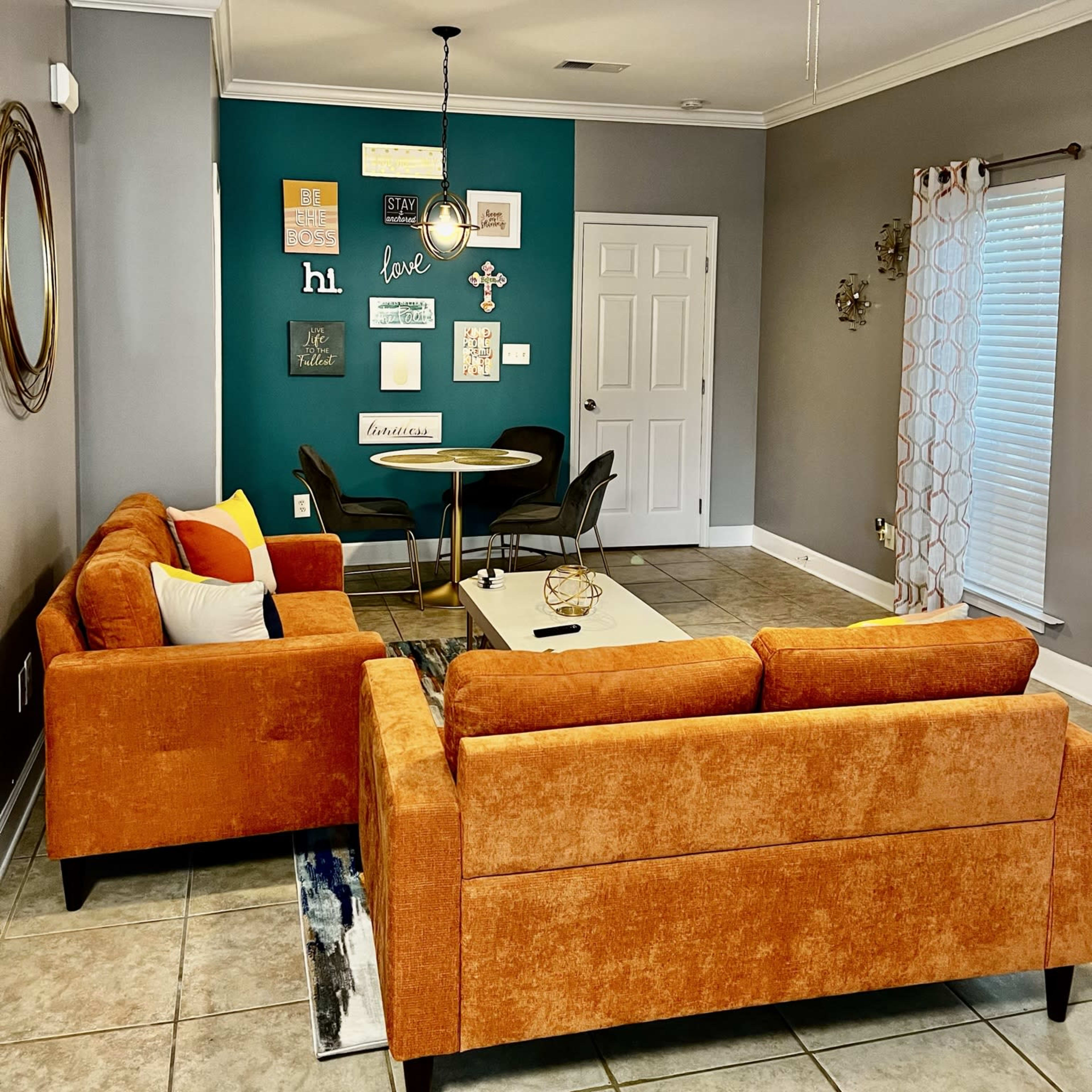 A living room with orange sofas facing a dining area, a teal accent wall adorned with framed artwork, and a ceiling fan overhead.