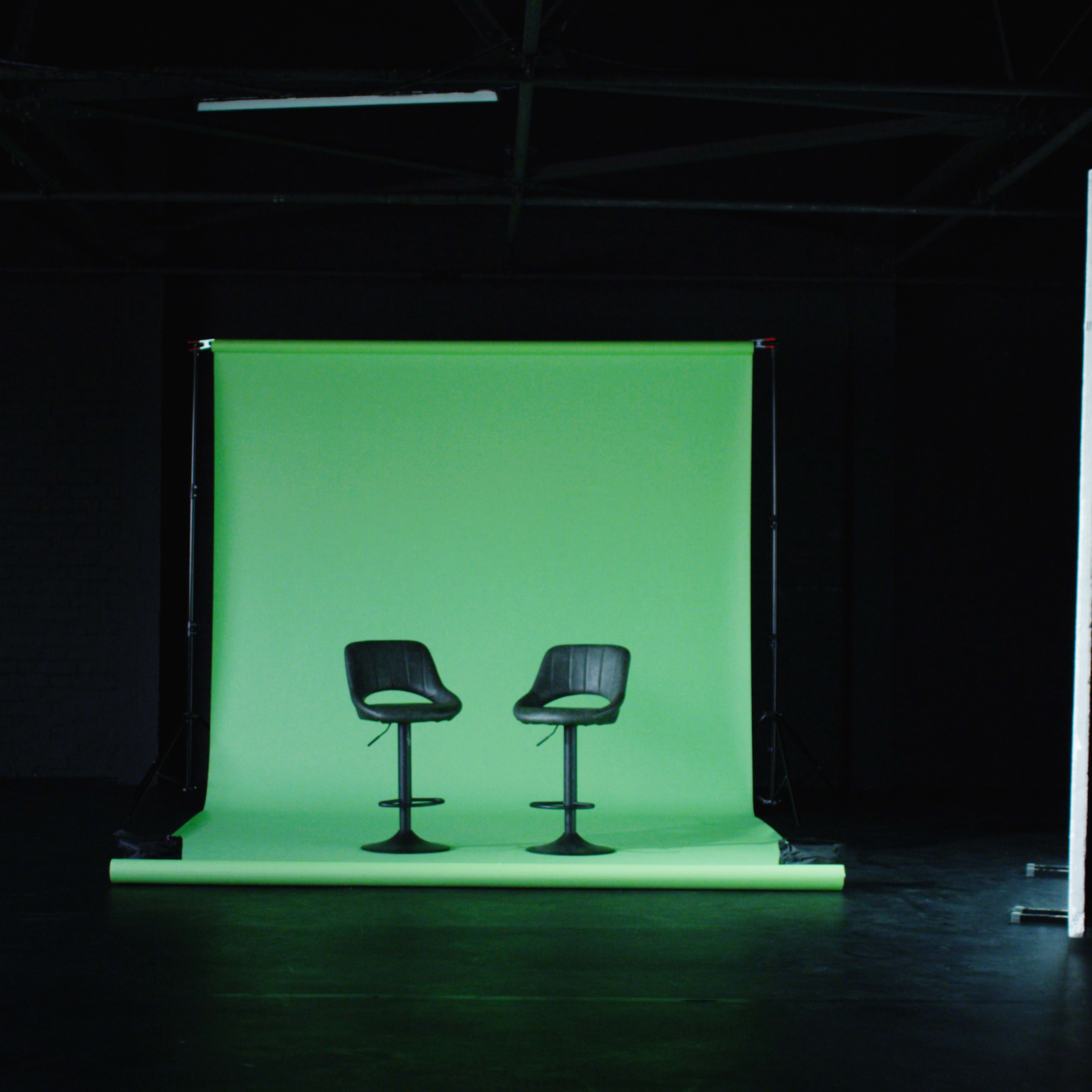 Two black bar stools positioned in front of a green backdrop inside a dimly lit studio.