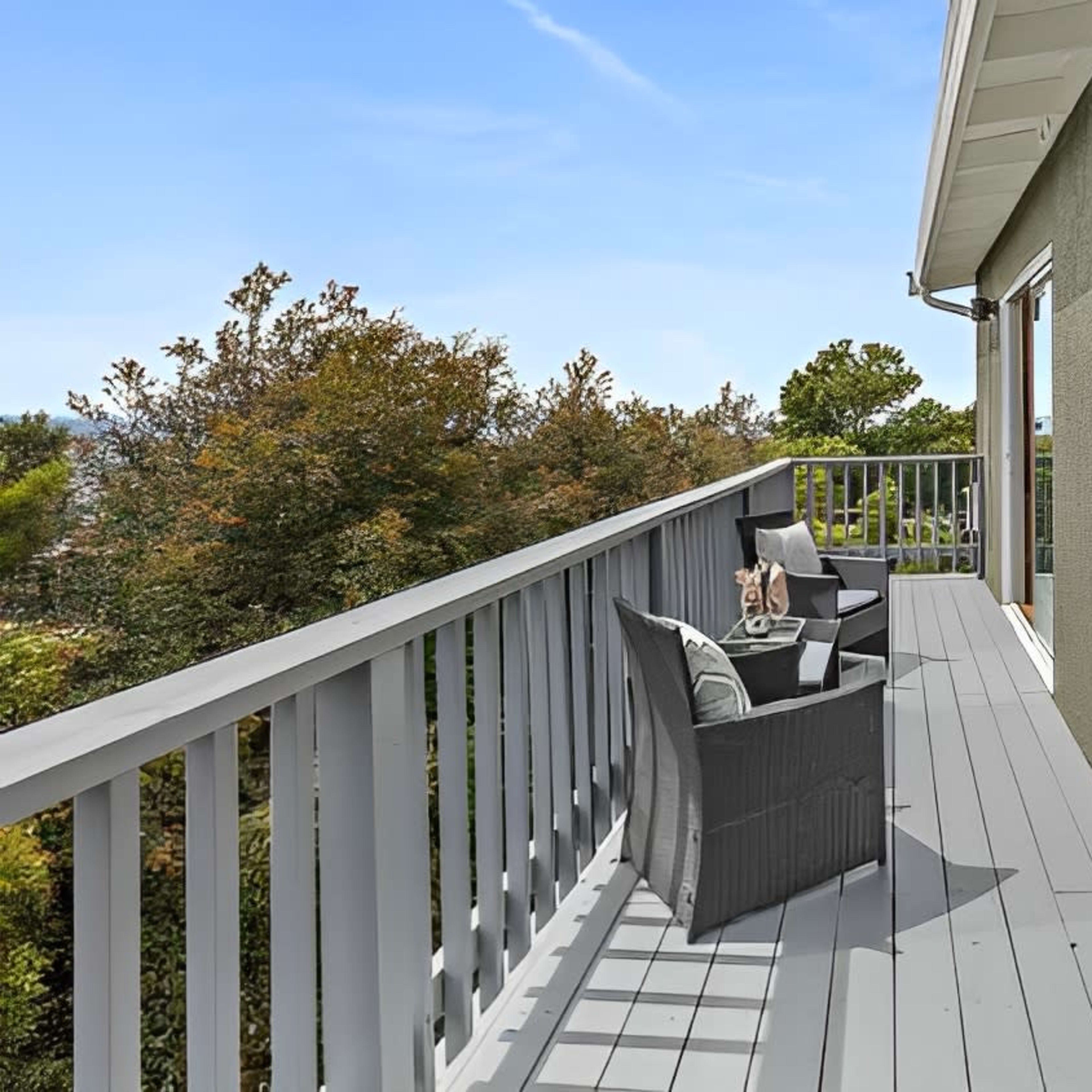 A balcony features two chairs and a small table, overlooking trees in a residential area.